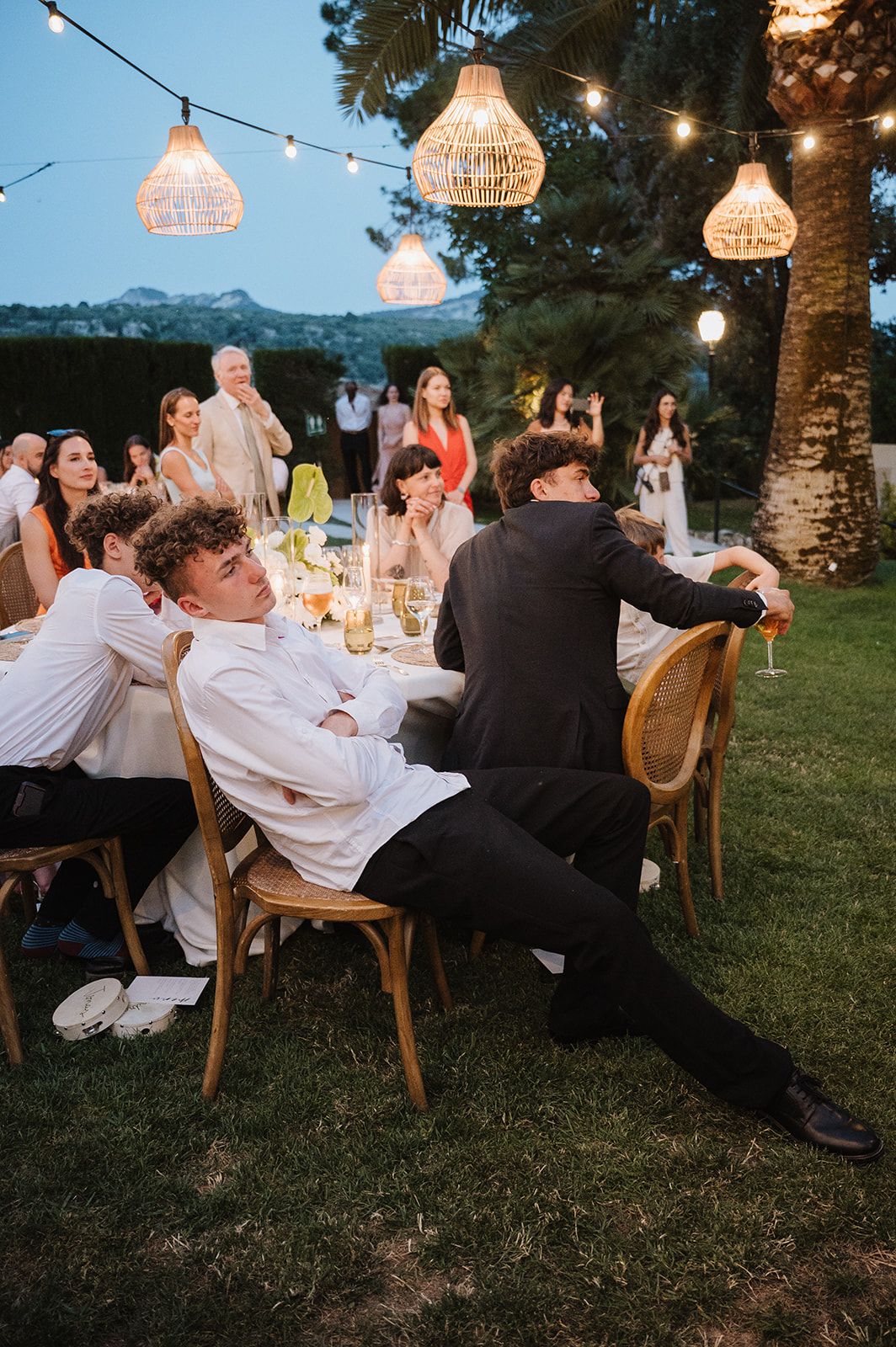 Young wedding guests relaxing at outdoor evening reception under warm string lights and wicker pendants.