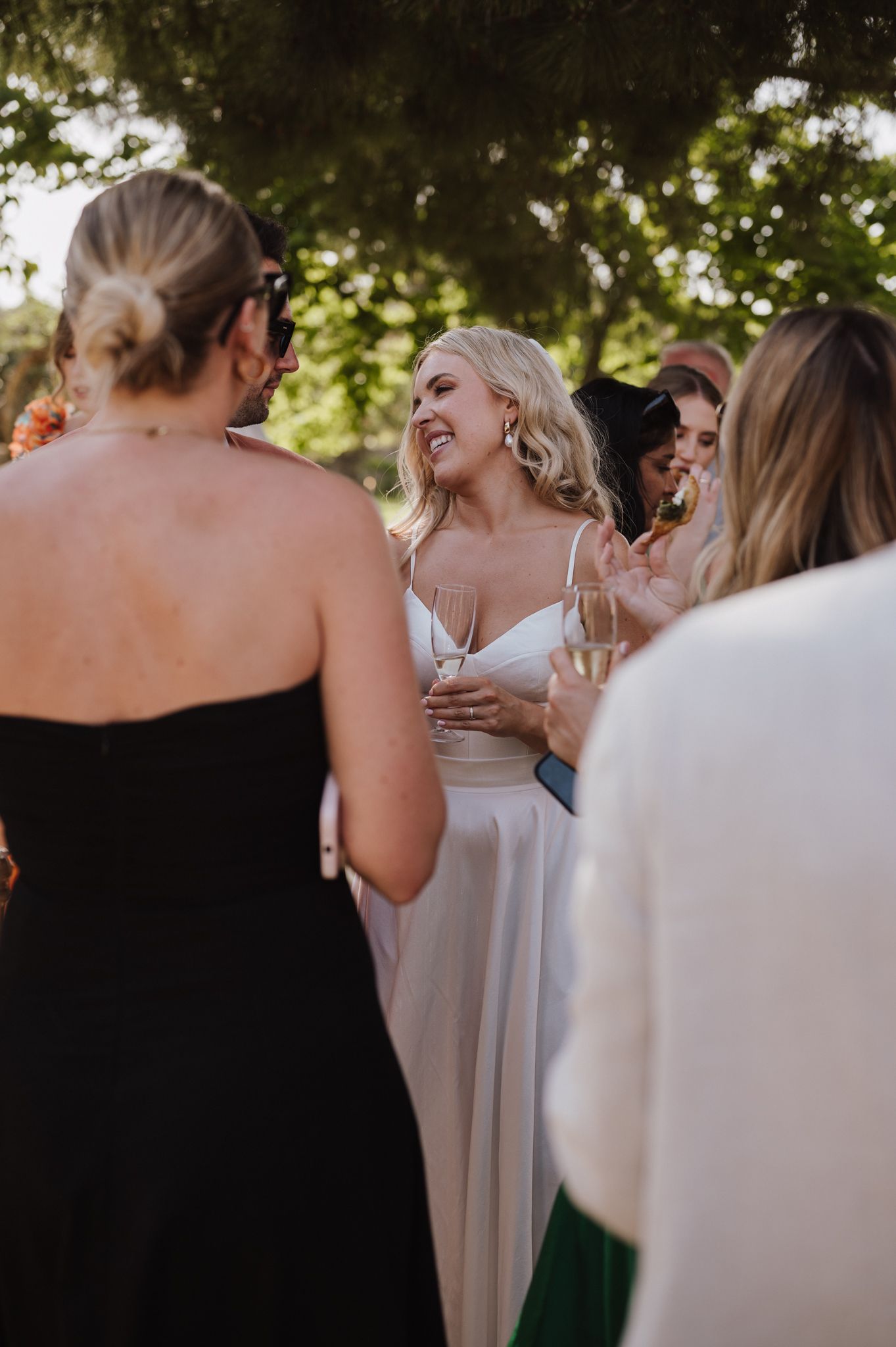 Smiling bride in white gown holds champagne while chatting with guests during outdoor wedding cocktail hour.
