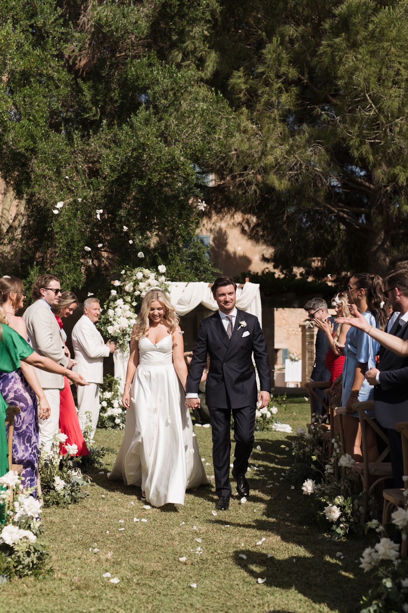 Bride and groom walk down outdoor wedding aisle lined with white floral arrangements and cheering guests.