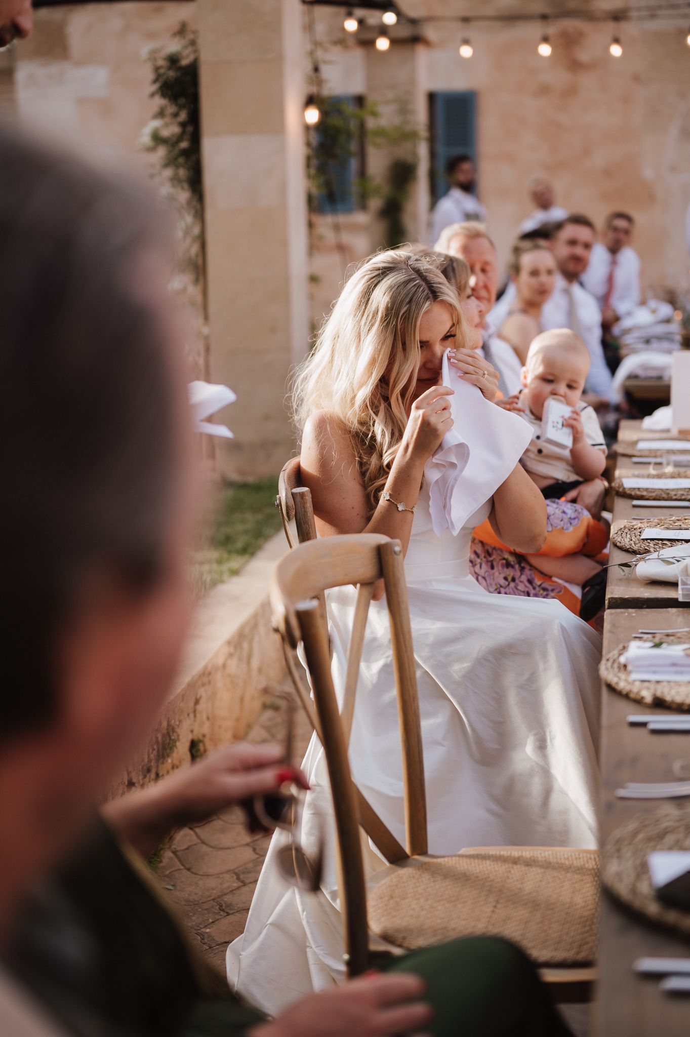 Bride in white gown holds baby at long outdoor wedding reception table with string lights overhead.