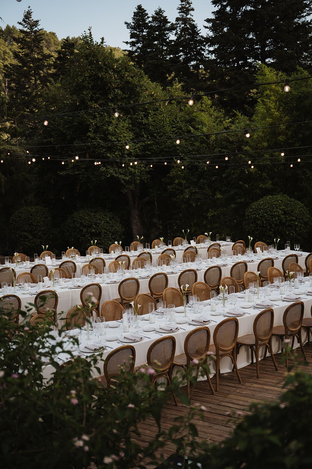 Elegant outdoor wedding reception with long tables, rattan chairs and string lights at dusk.