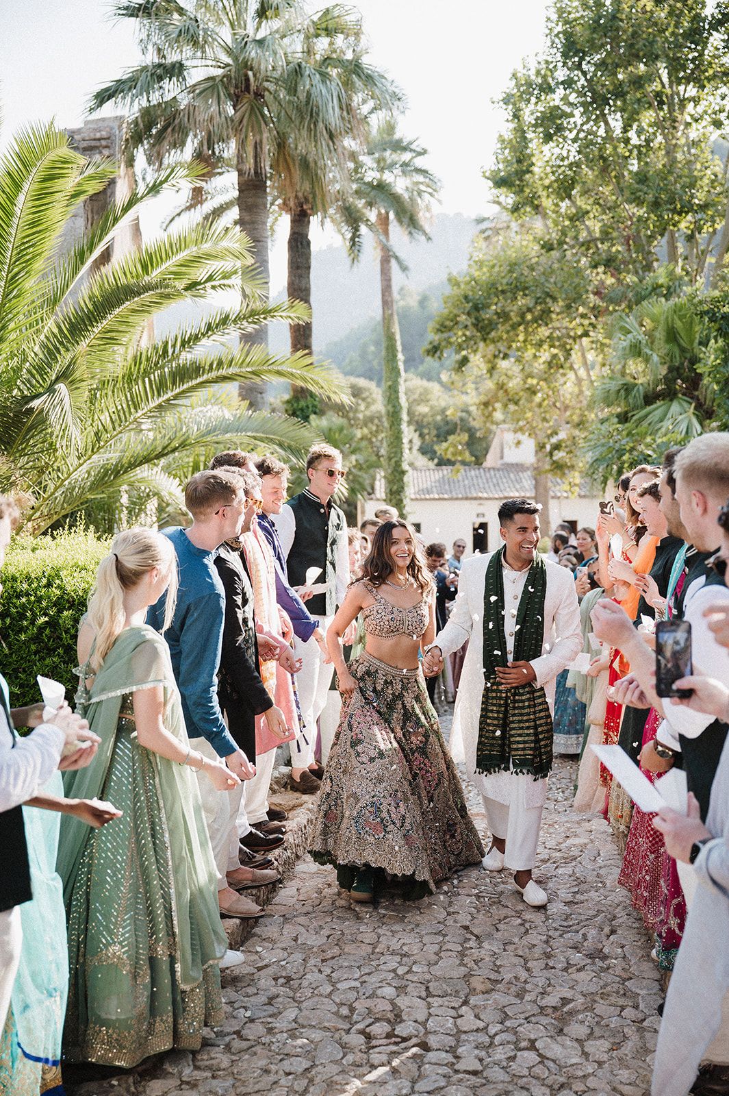 Bride in ornate lehenga and groom walking through cheering guests at outdoor Indian wedding.