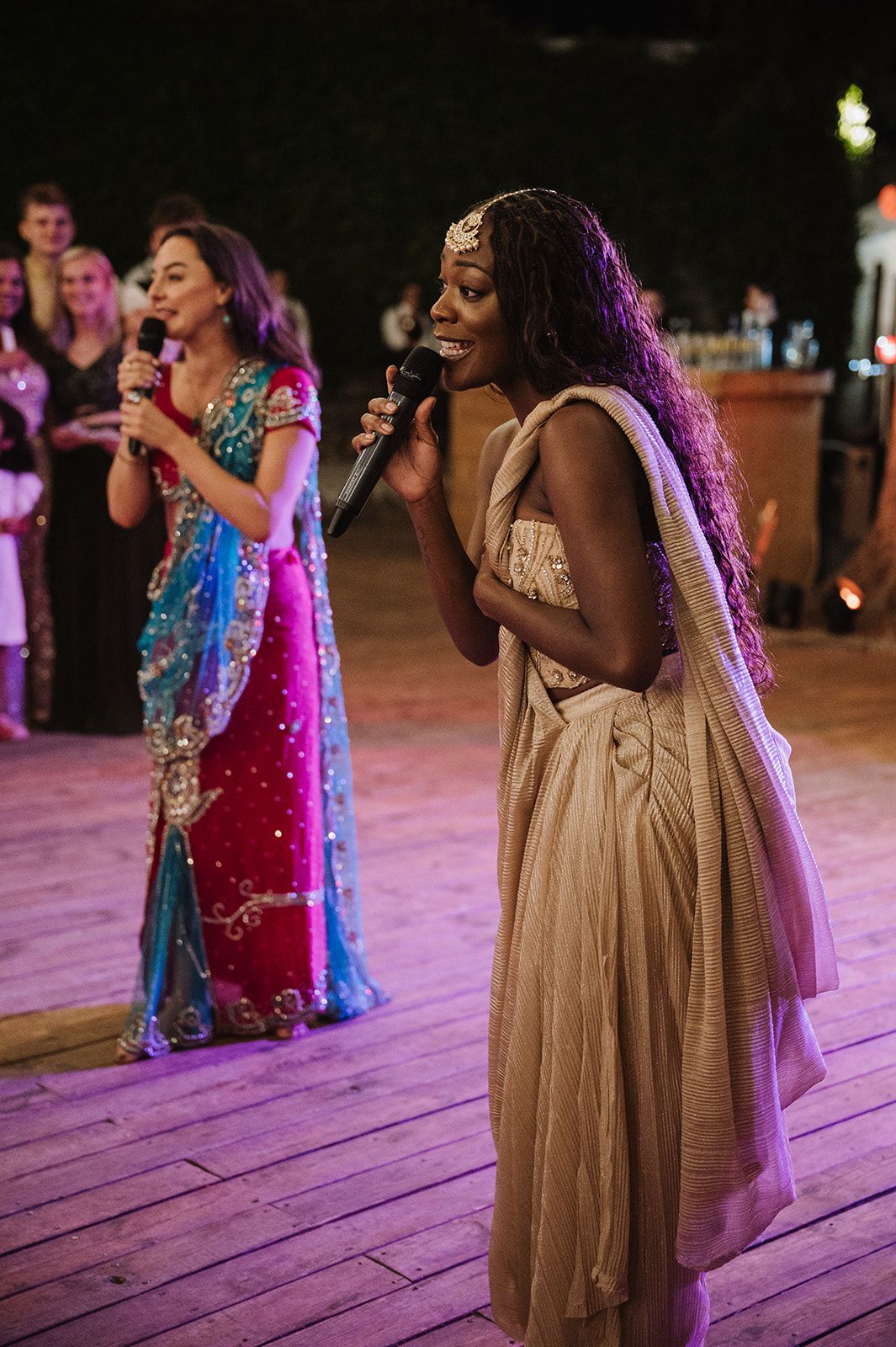 Women performing with microphones in elegant saree and gown at Indian wedding reception celebration.