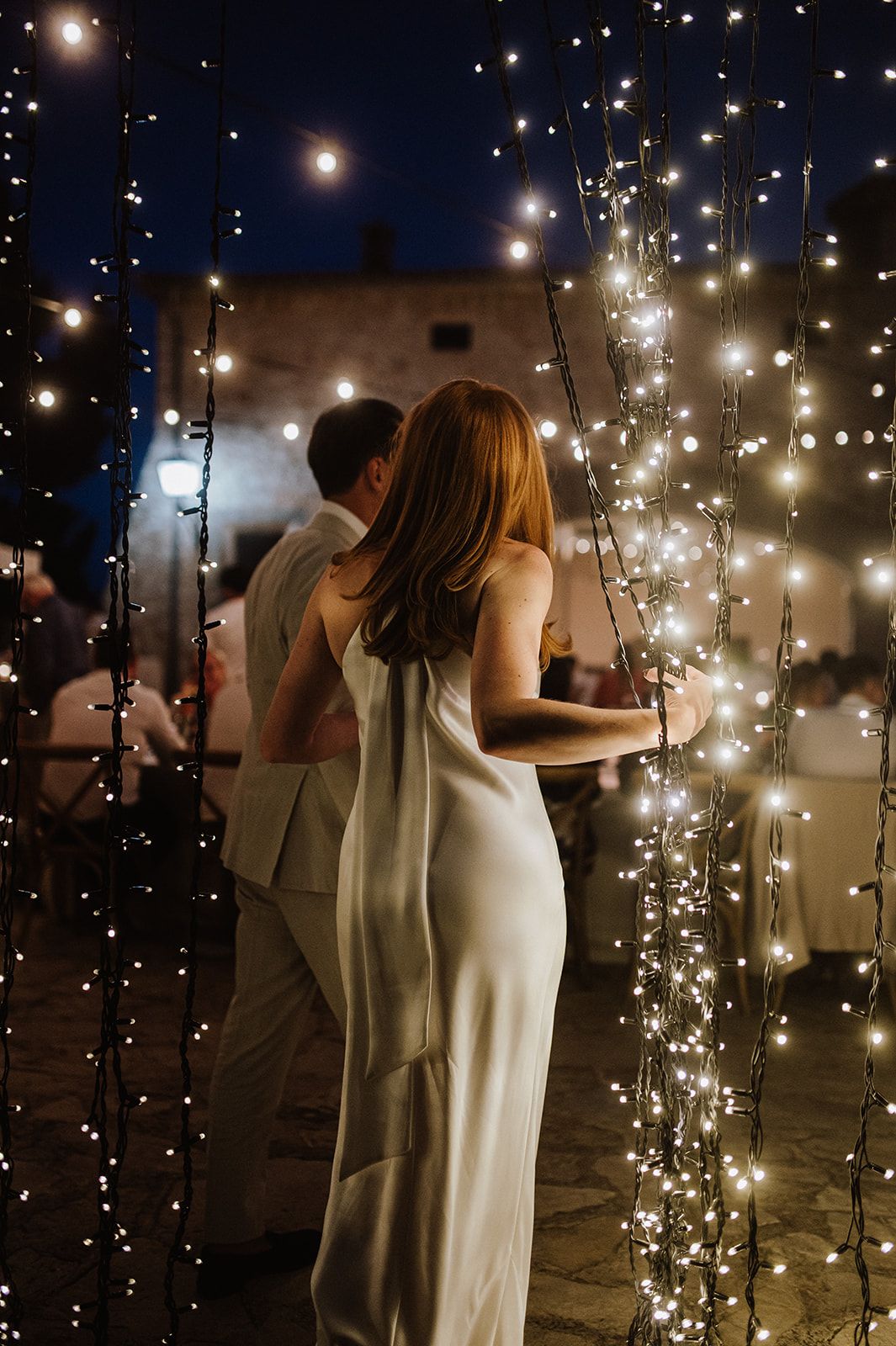 Bride and groom share a romantic moment surrounded by cascading fairy lights at an outdoor evening wedding reception.