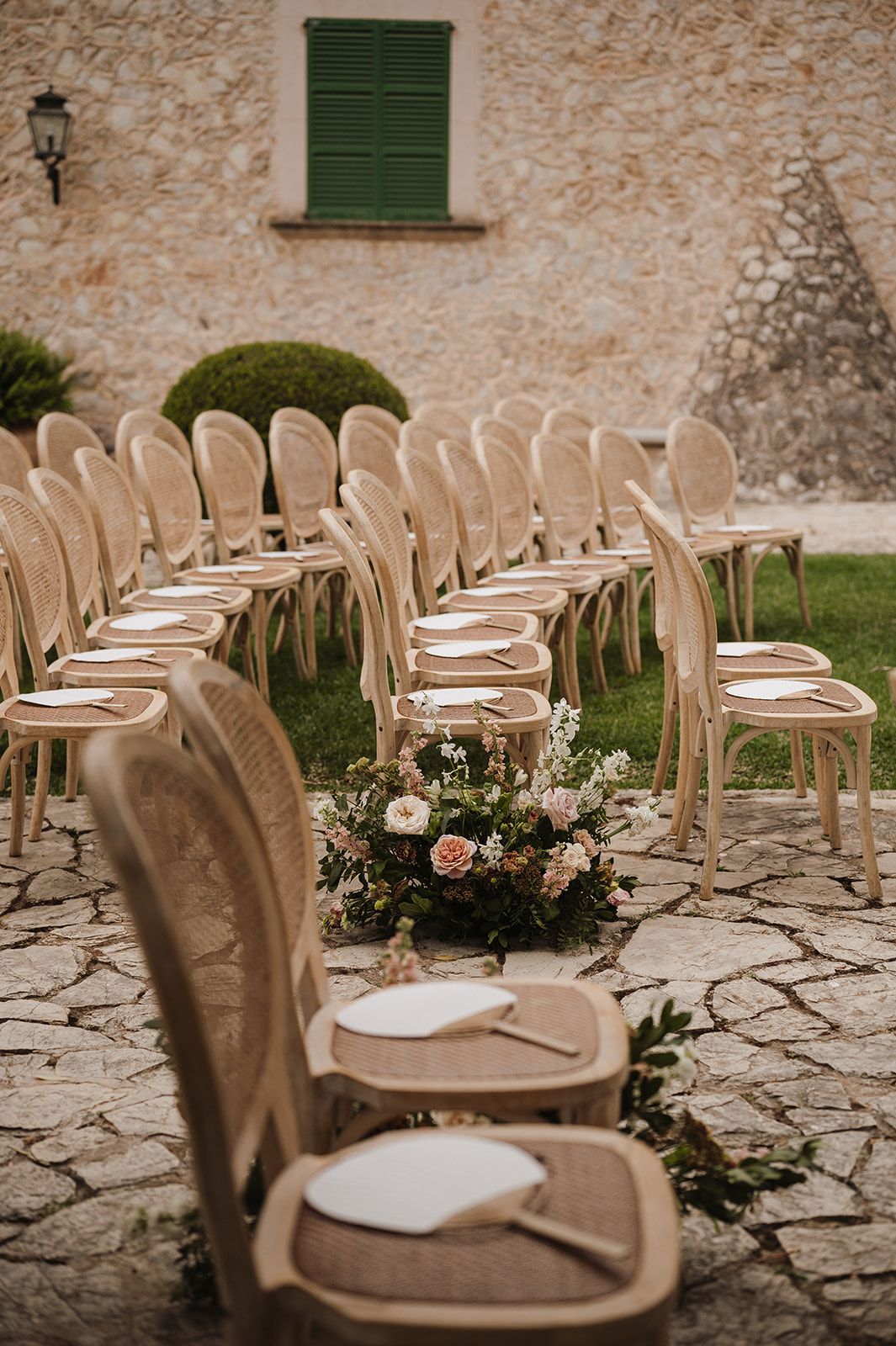 Rows of rattan chairs with white programs and floral aisle decor at outdoor wedding ceremony.