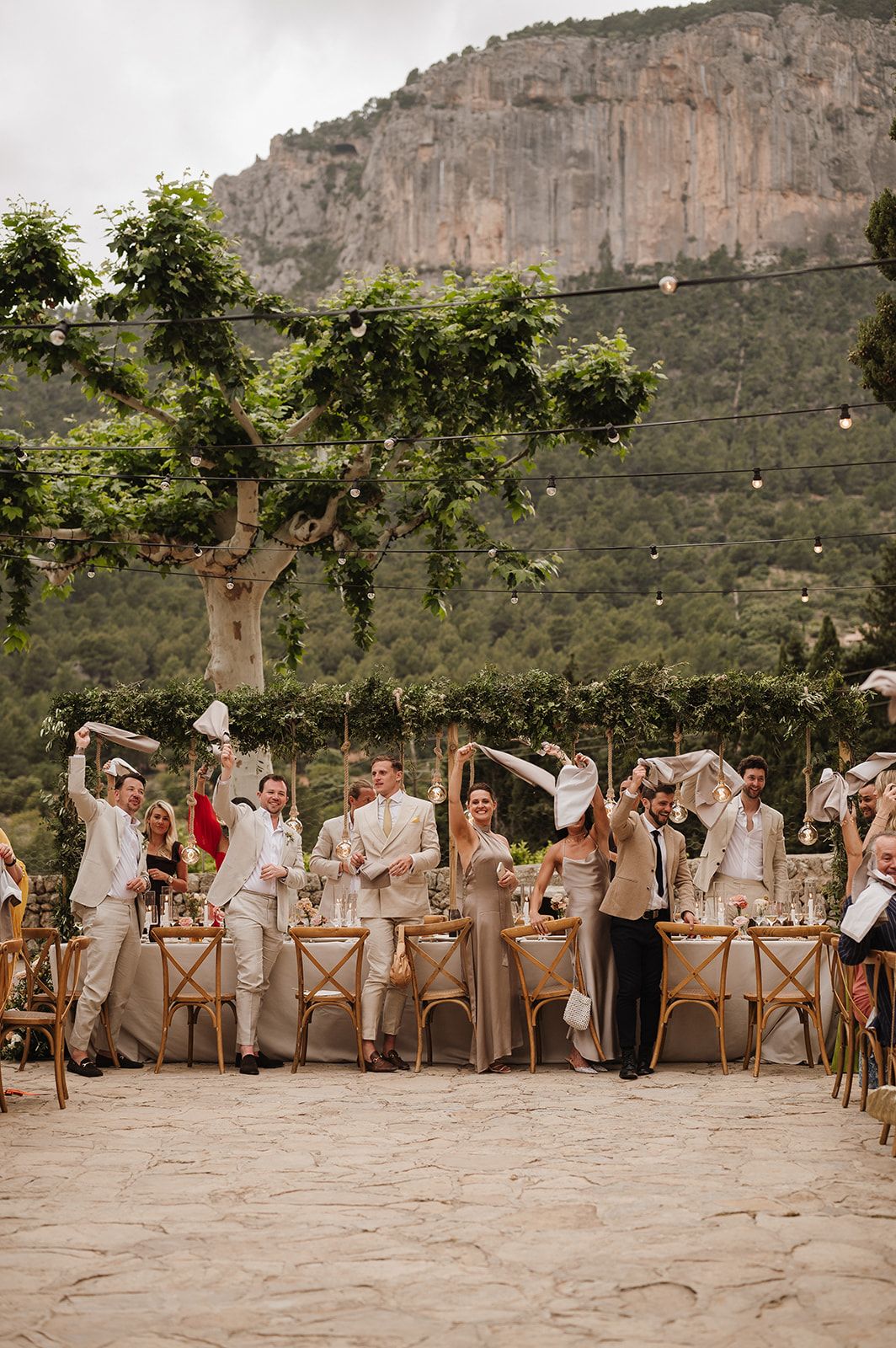 Wedding party waves napkins joyfully at outdoor reception with dramatic mountain backdrop.