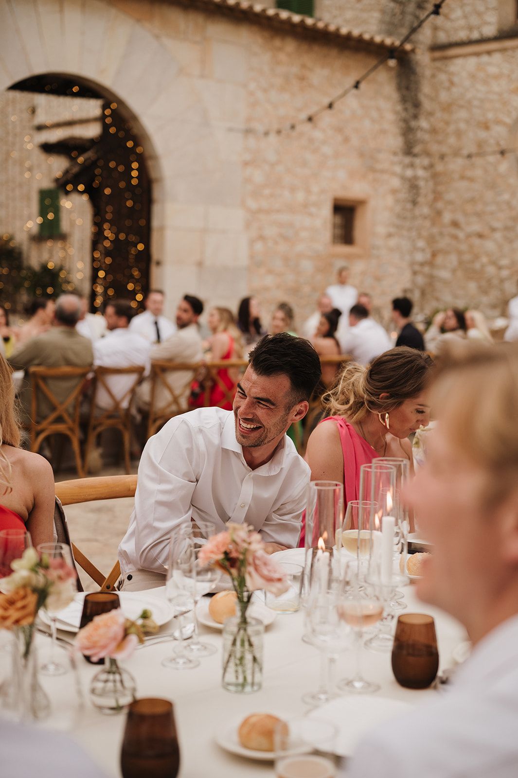 Smiling wedding guest in white shirt laughs at a candlelit outdoor reception table at a rustic stone venue.