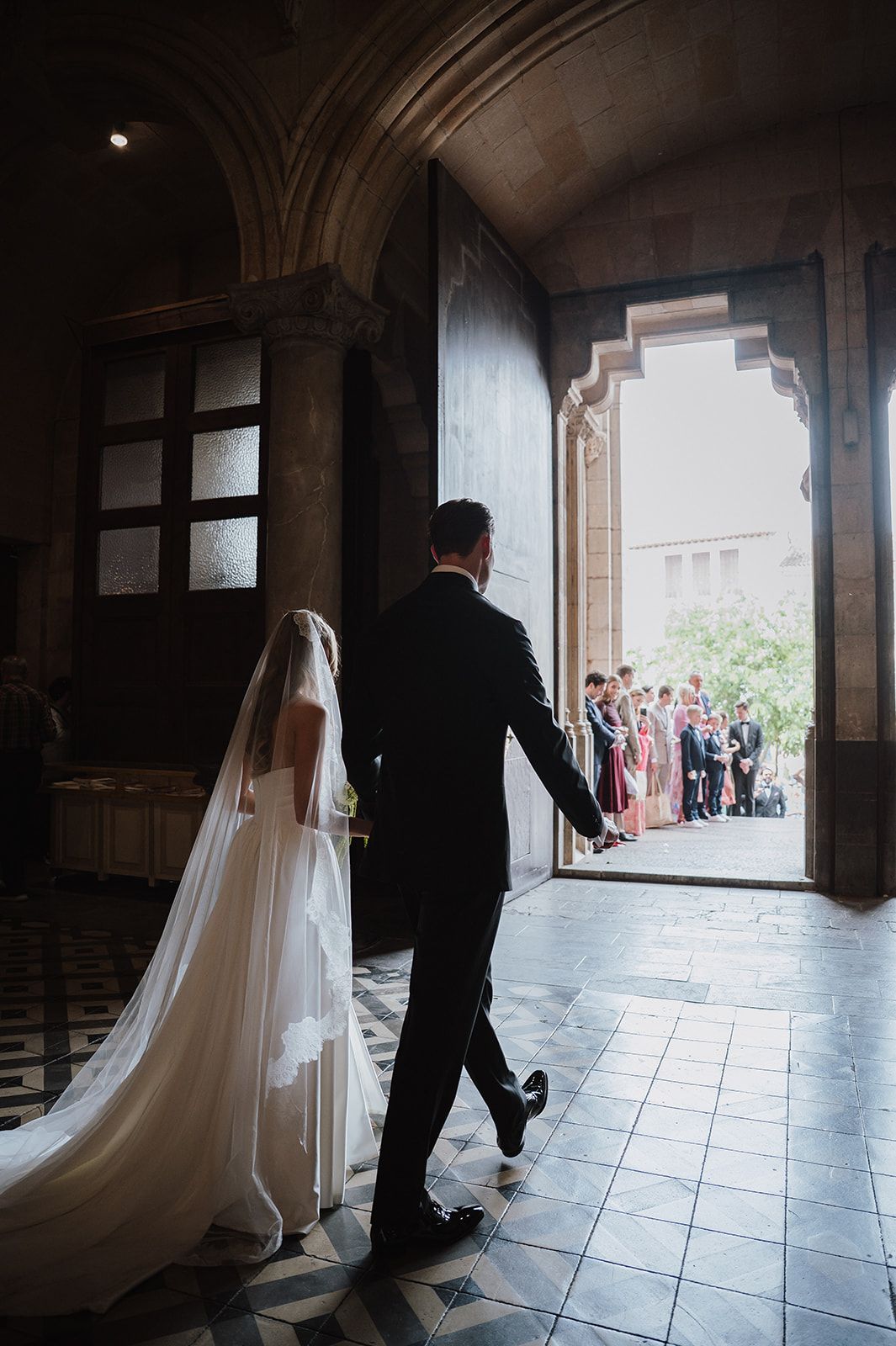 Bride in flowing white gown with cathedral veil walks with groom toward grand arched church entrance doors.