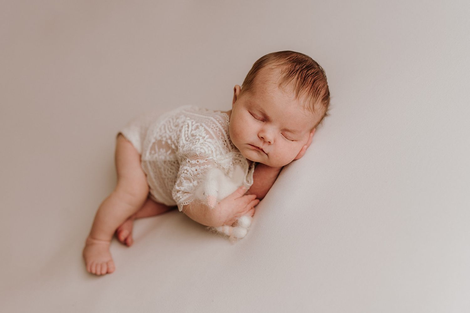Sleeping newborn baby in white lace romper posed on soft cream backdrop for professional newborn photography session.
