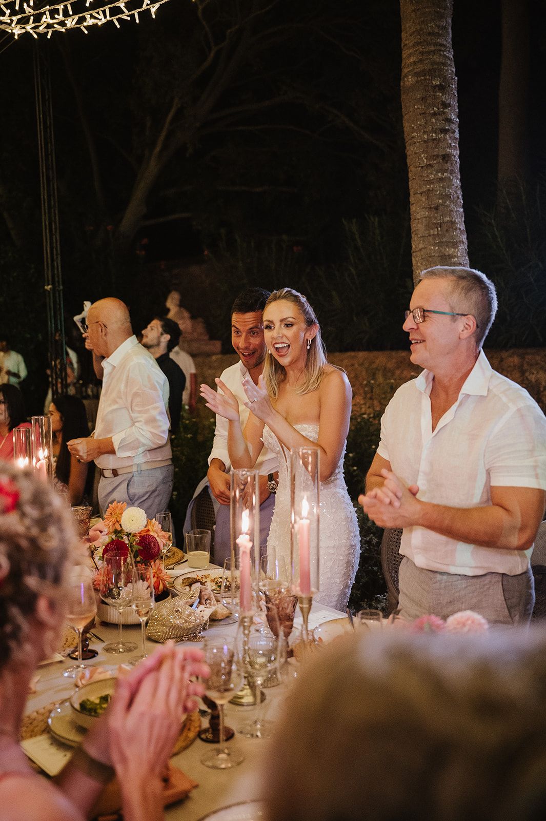 Bride in white gown laughs and claps at candlelit outdoor wedding reception dinner table.