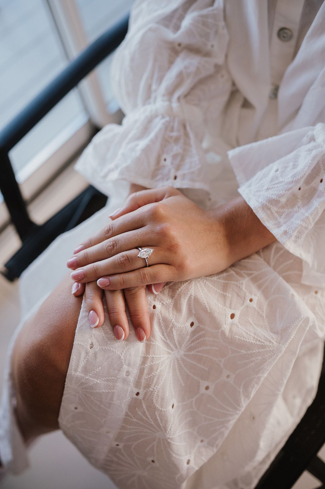 Woman in white eyelet dress displaying a sparkling diamond engagement ring on her finger while seated.