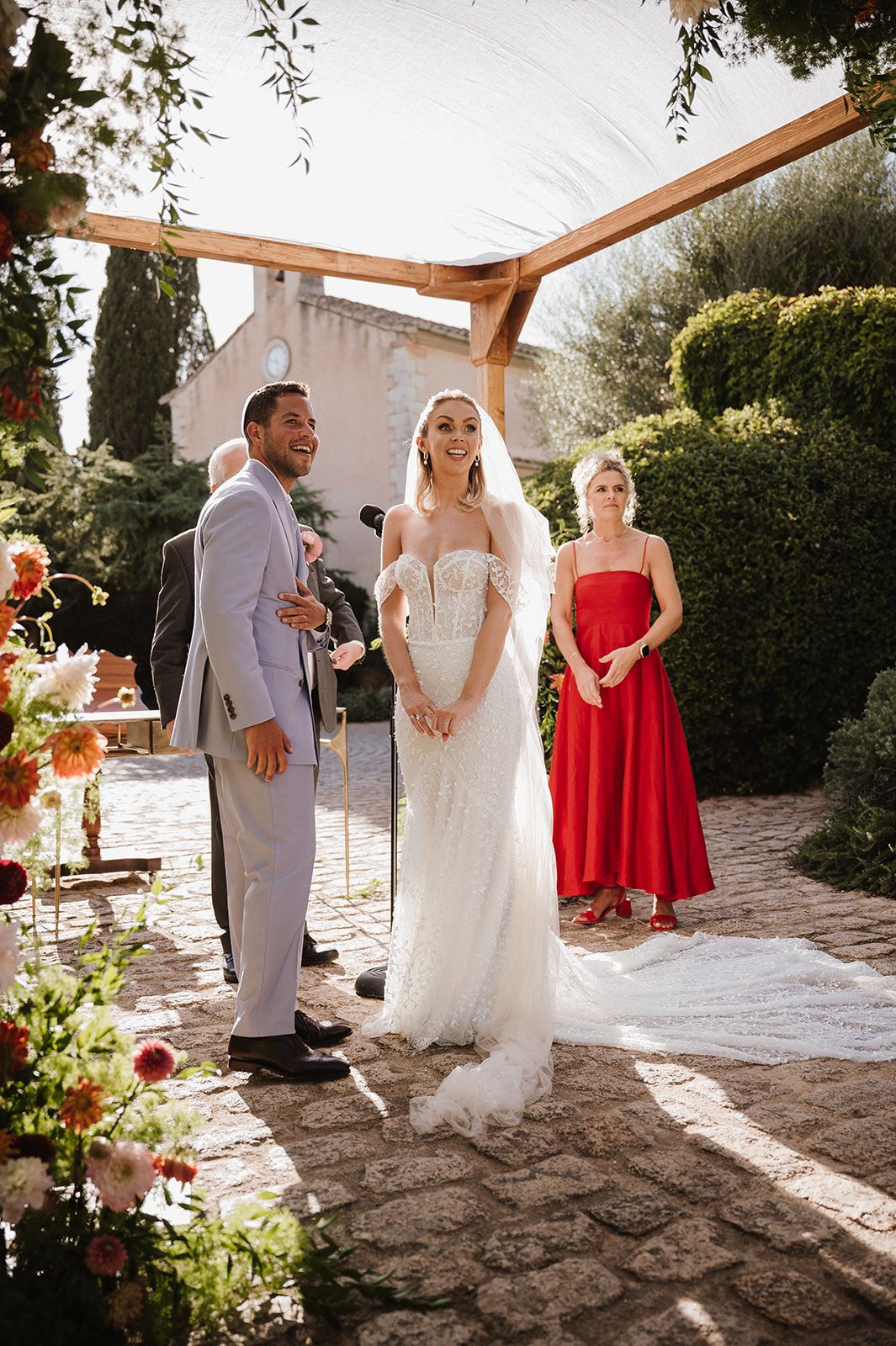 Smiling bride in feathered lace gown and groom in grey suit standing under a wooden floral chuppah outdoors.