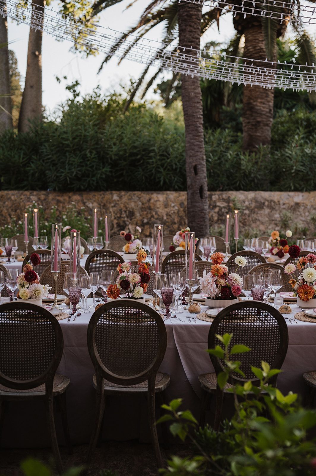 Elegant outdoor wedding reception table set with pink candles, floral centerpieces, and rattan chairs at dusk.