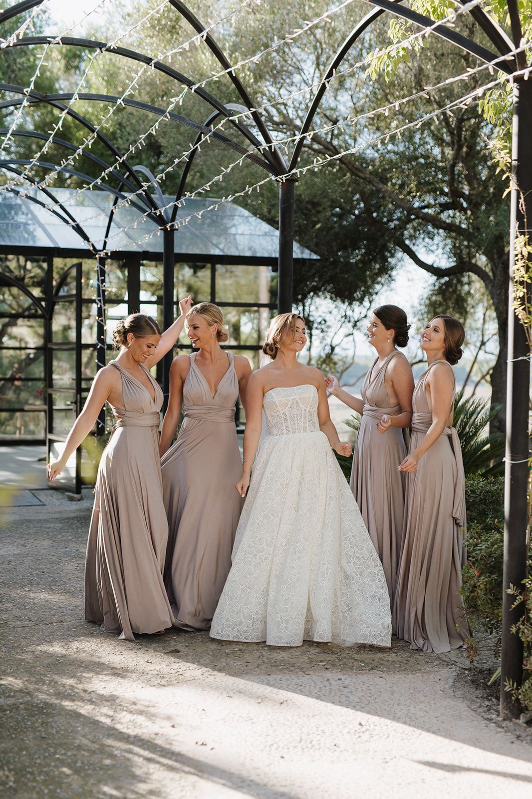 Bride in lace gown with four bridesmaids in taupe dresses under garden pergola.
