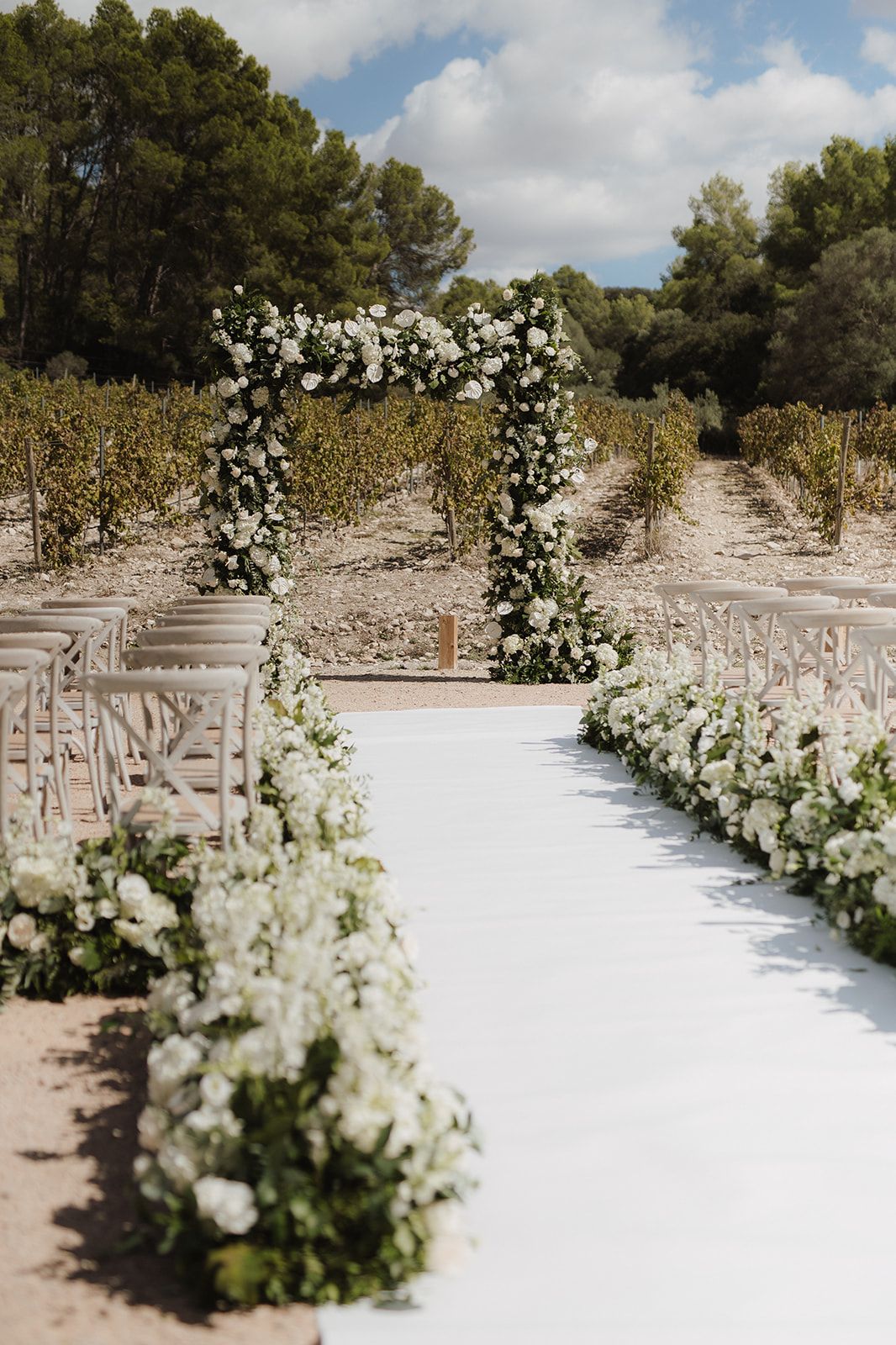 Elegant outdoor wedding aisle with white floral arch and flower-lined white runner in a vineyard setting.