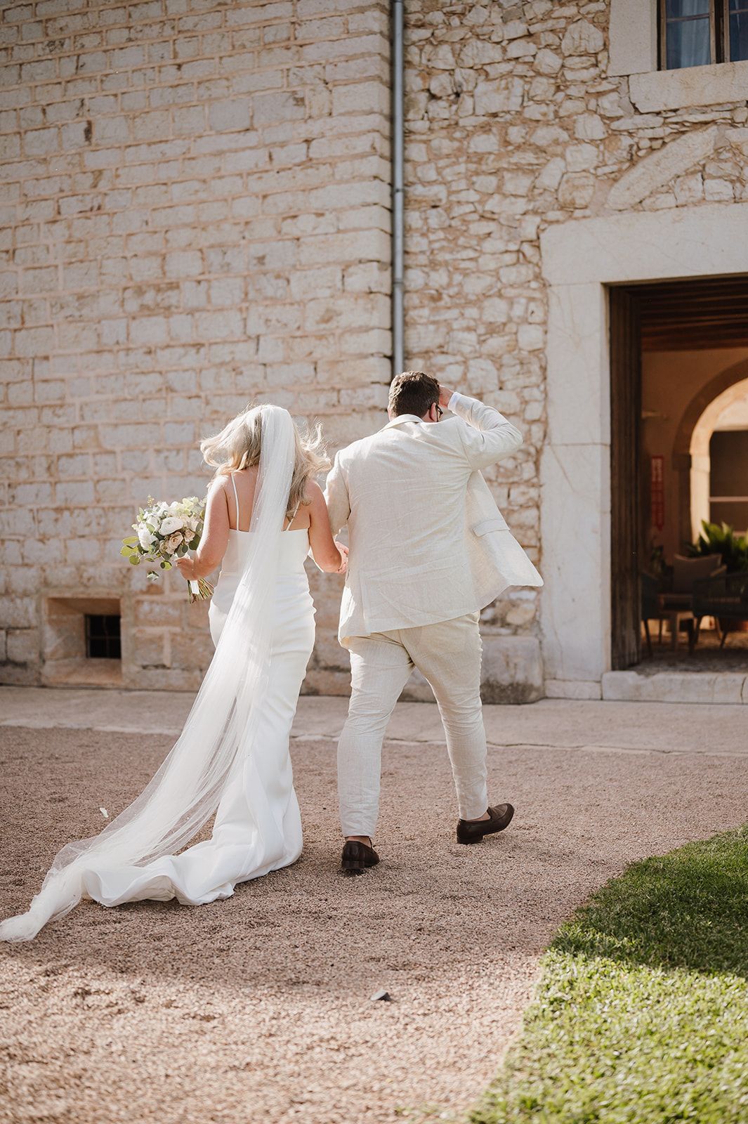 Bride and groom walking away from stone building, bride holding bouquet with cathedral veil trailing.