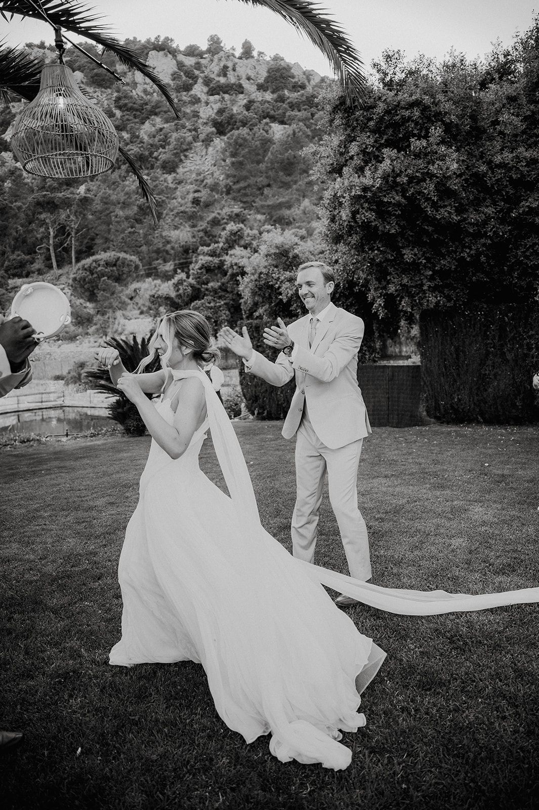 Black and white photo of bride and groom dancing playfully on garden lawn at outdoor wedding reception.