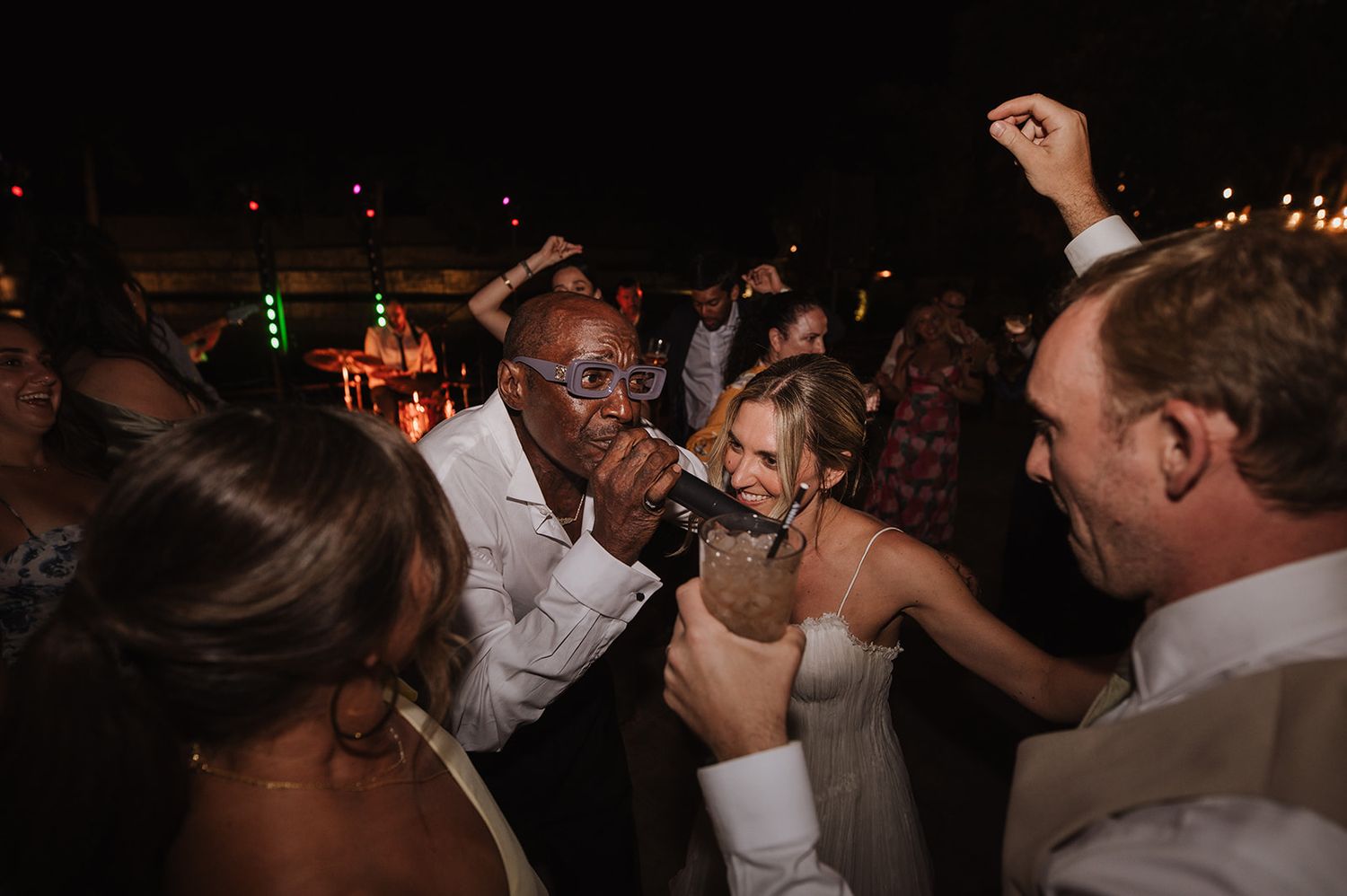 Guests dancing and singing at a lively outdoor wedding reception party at night.