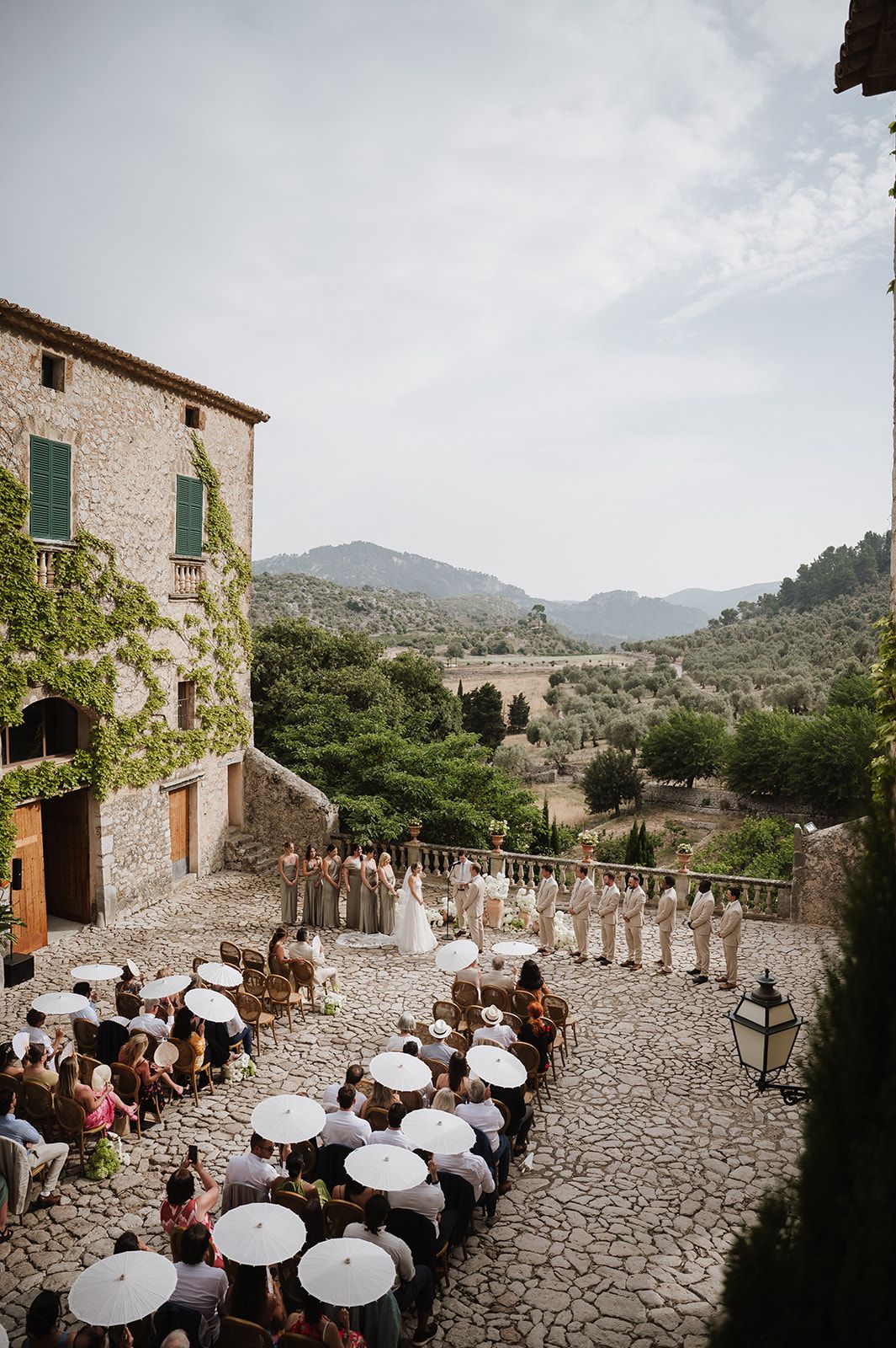 Outdoor wedding ceremony at a stone villa with mountain views, white umbrellas, and guests seated on cobblestones.