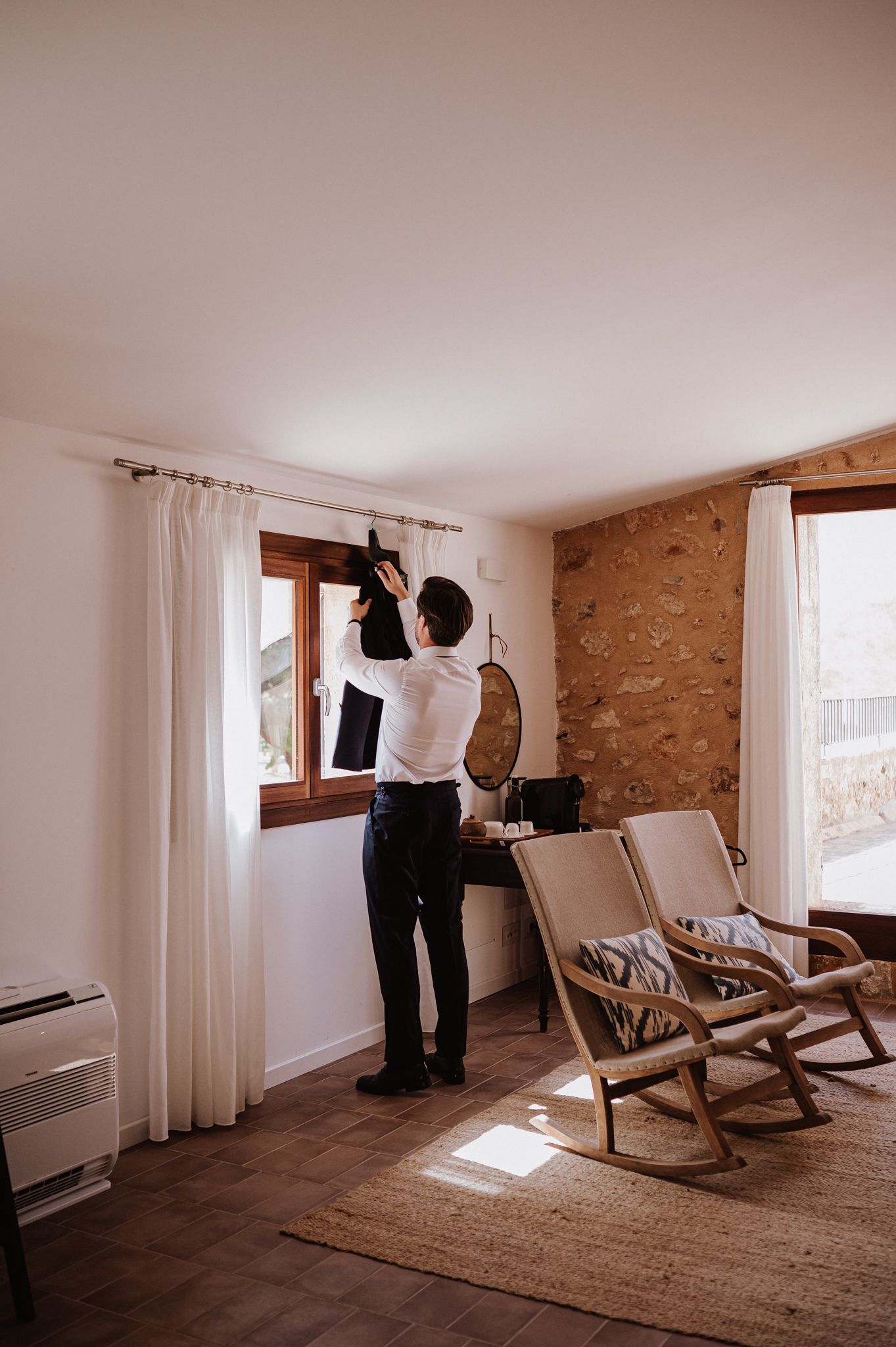 Groom in white shirt reaching up to retrieve his black suit jacket before the ceremony.
