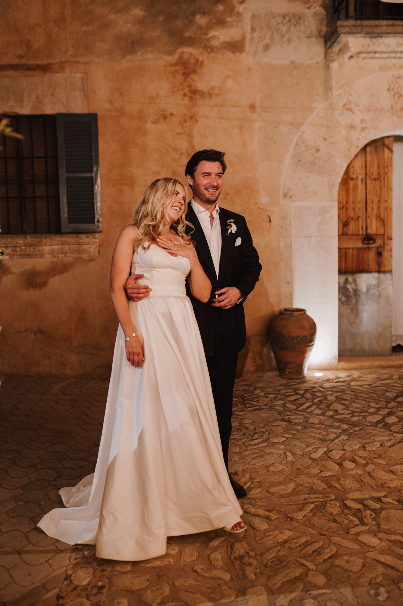 Bride and groom laugh together on cobblestone courtyard at night with warm stone wall backdrop.