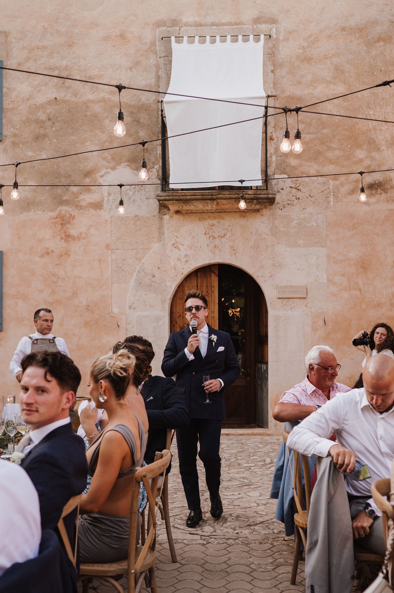 Groom in navy suit gives speech with microphone at outdoor wedding reception in rustic stone courtyard with string lights.