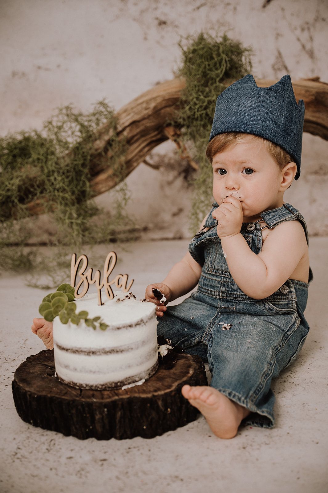 Baby boy in denim overalls and crown hat smashing first birthday cake on wood slice with greenery backdrop.