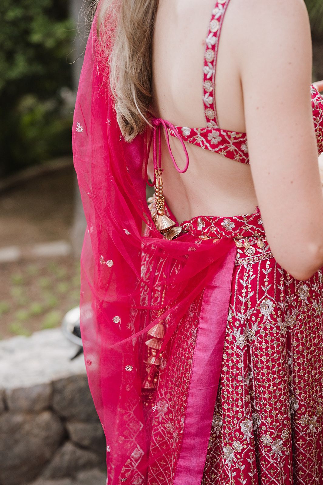 Back view of woman in vibrant pink lehenga with gold tassel embellishments at Indian wedding.