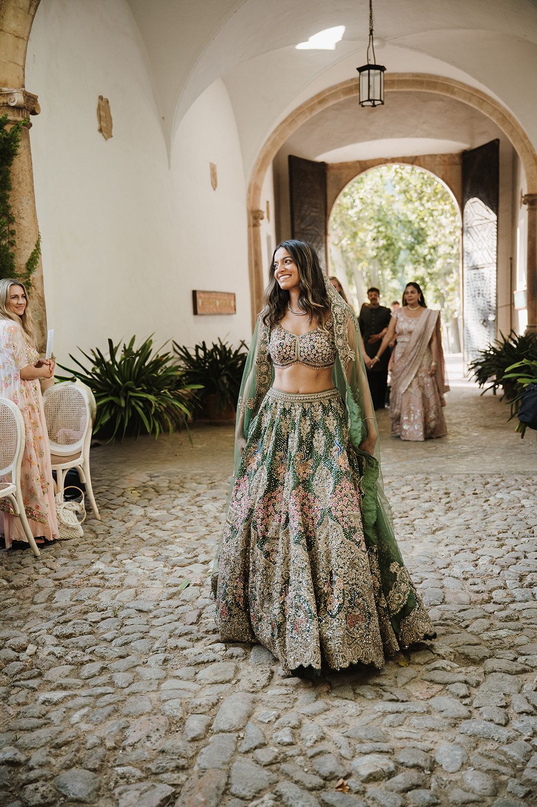 Smiling bride in green embroidered lehenga walking through stone archway at Indian wedding.