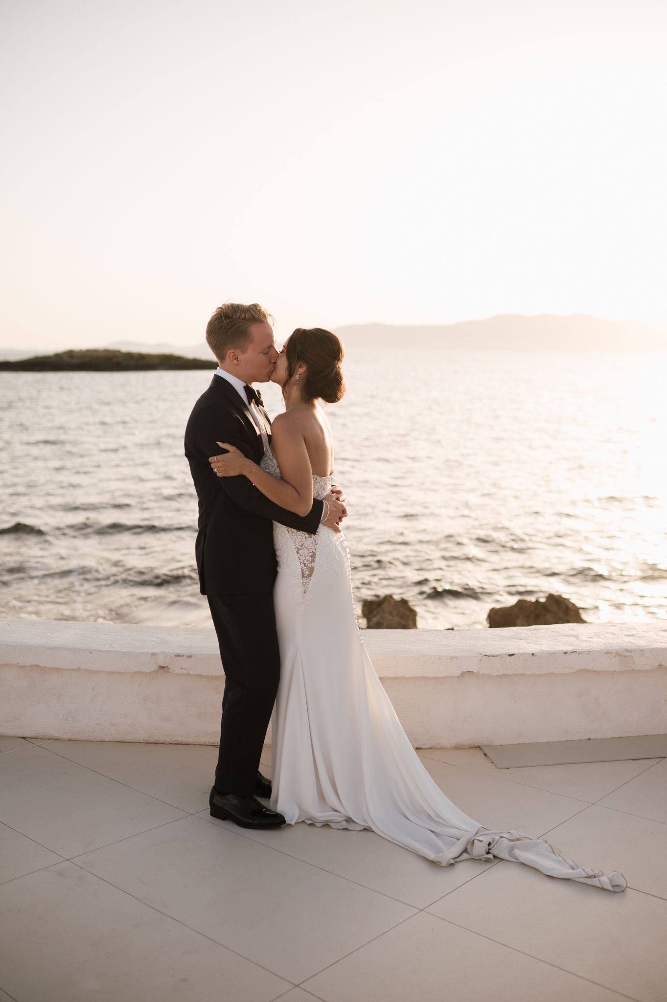 Bride and groom share a sunset kiss on a seaside terrace, brides elegant gown trailing behind her.