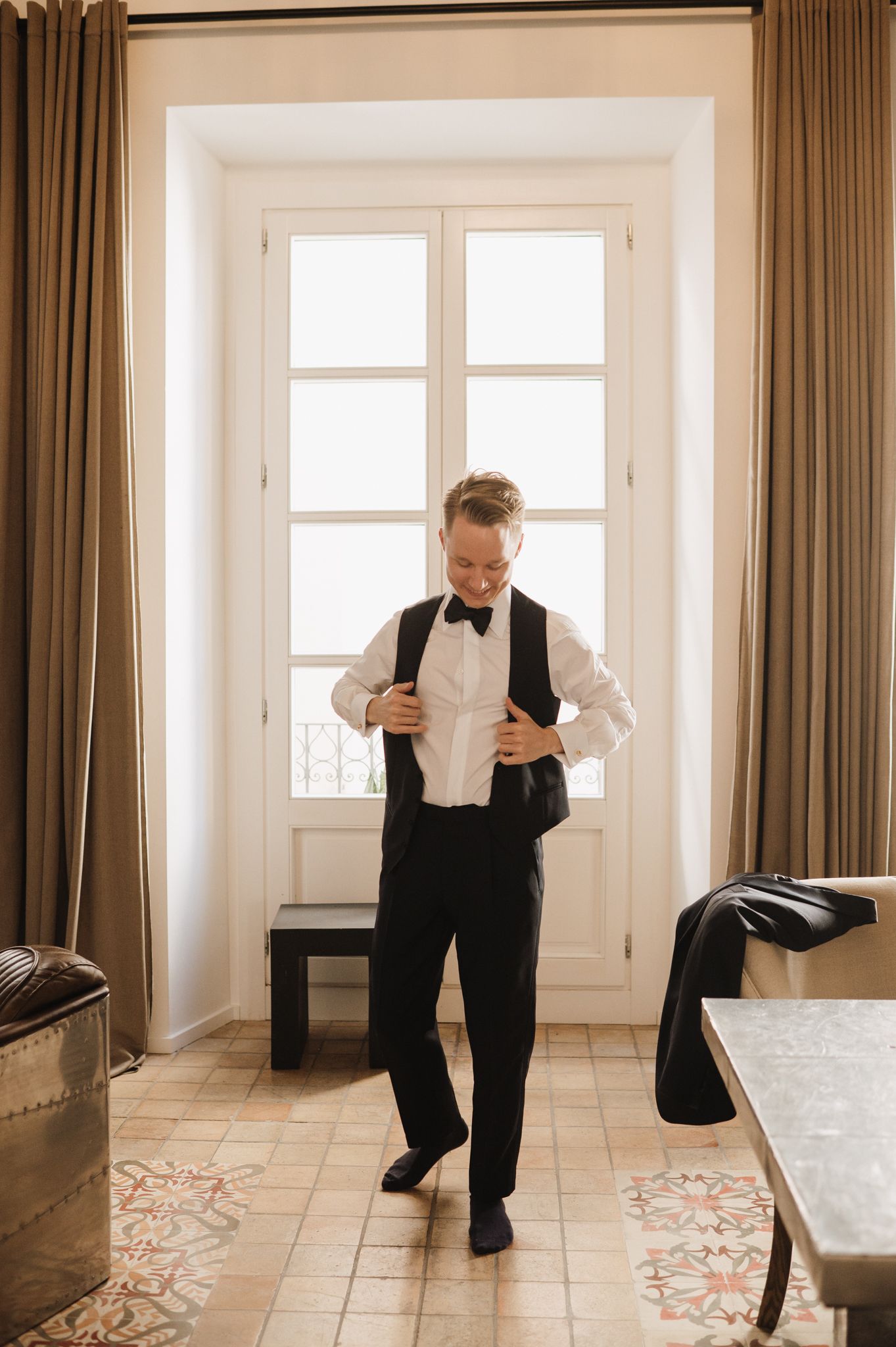 Groom in black tuxedo vest and bow tie adjusting his jacket while getting ready indoors near a bright window.