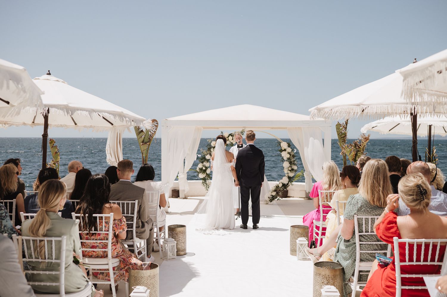 Outdoor seaside wedding ceremony with guests seated under white umbrellas on a sunny day.