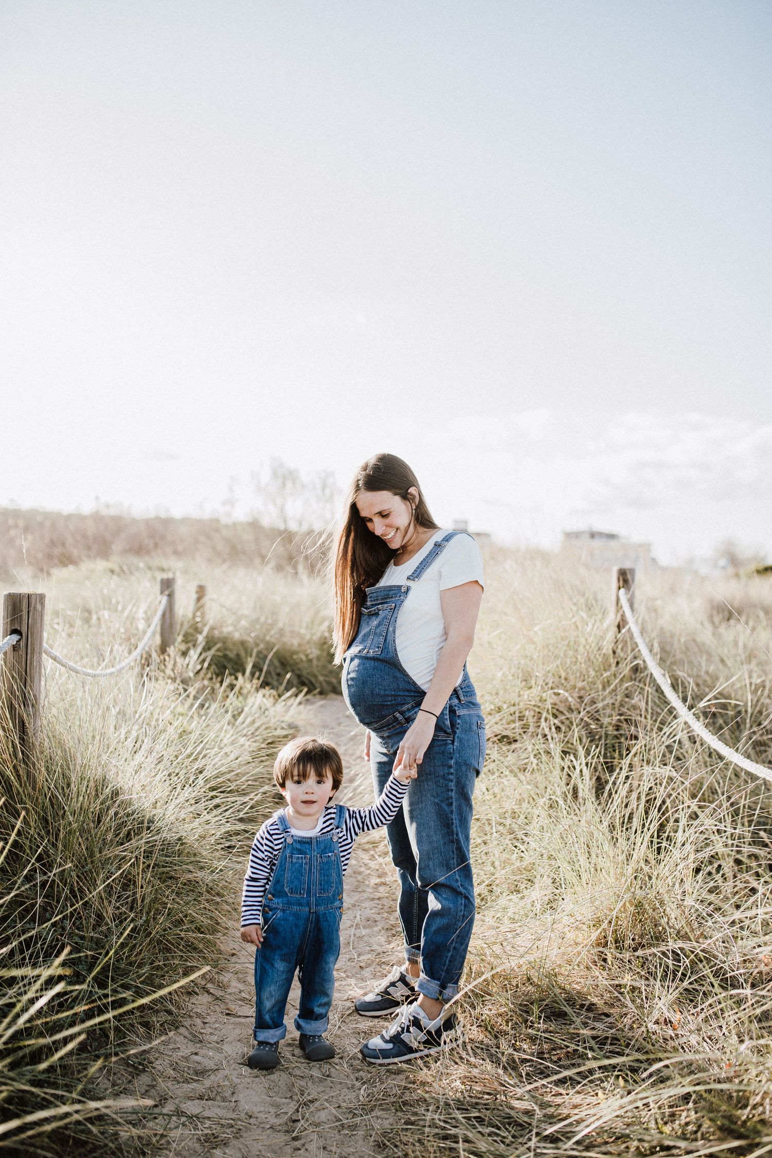 Pregnant woman in denim overalls holds toddler's hand on sandy path surrounded by tall grass.