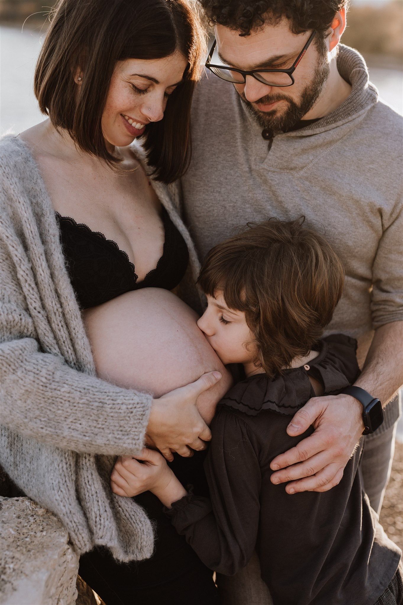 Pregnant woman in black bralette and grey cardigan with partner and child kissing baby bump outdoors.