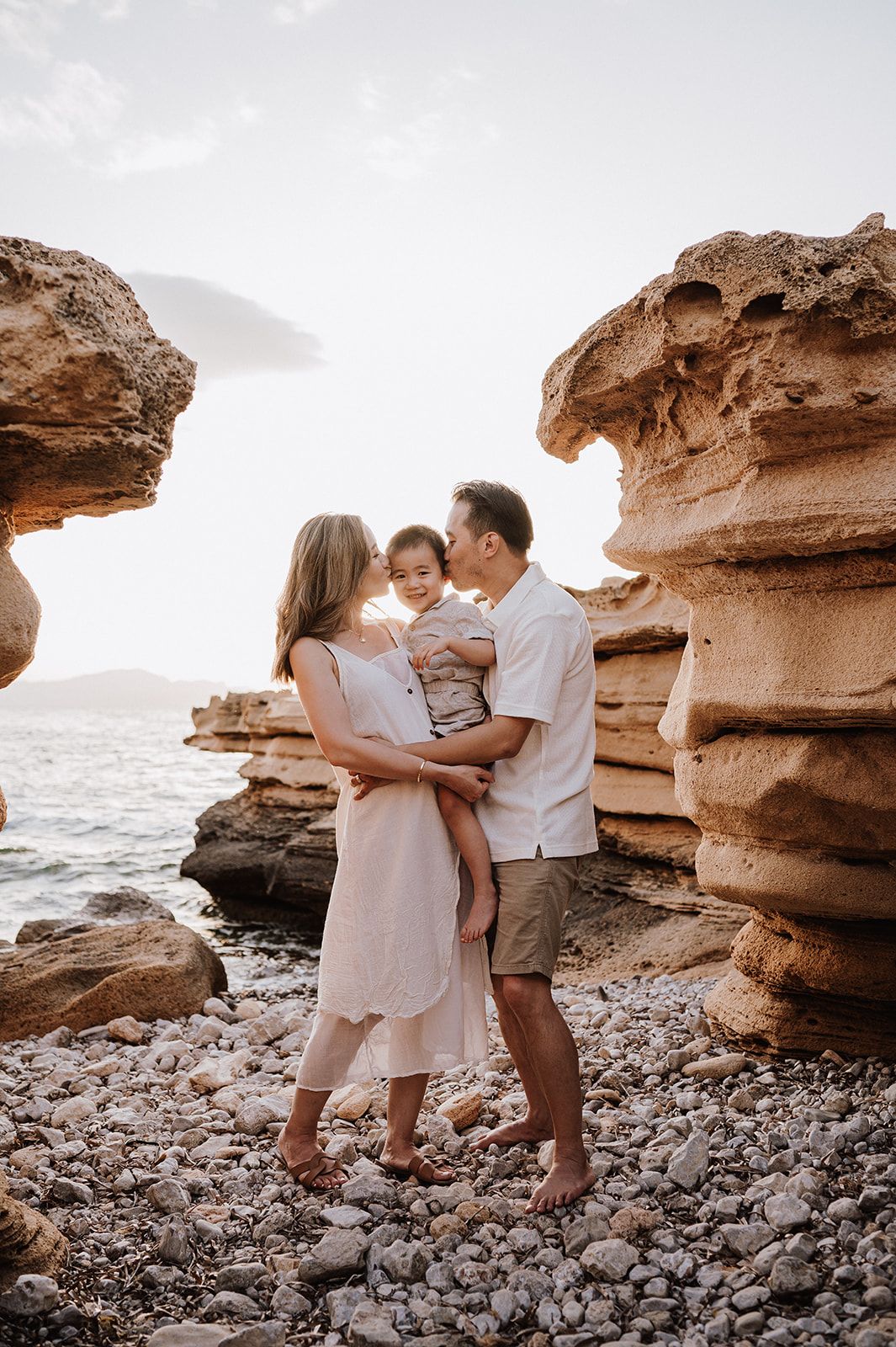 Family of three posing among golden rocky cliffs on a pebble beach at sunset.