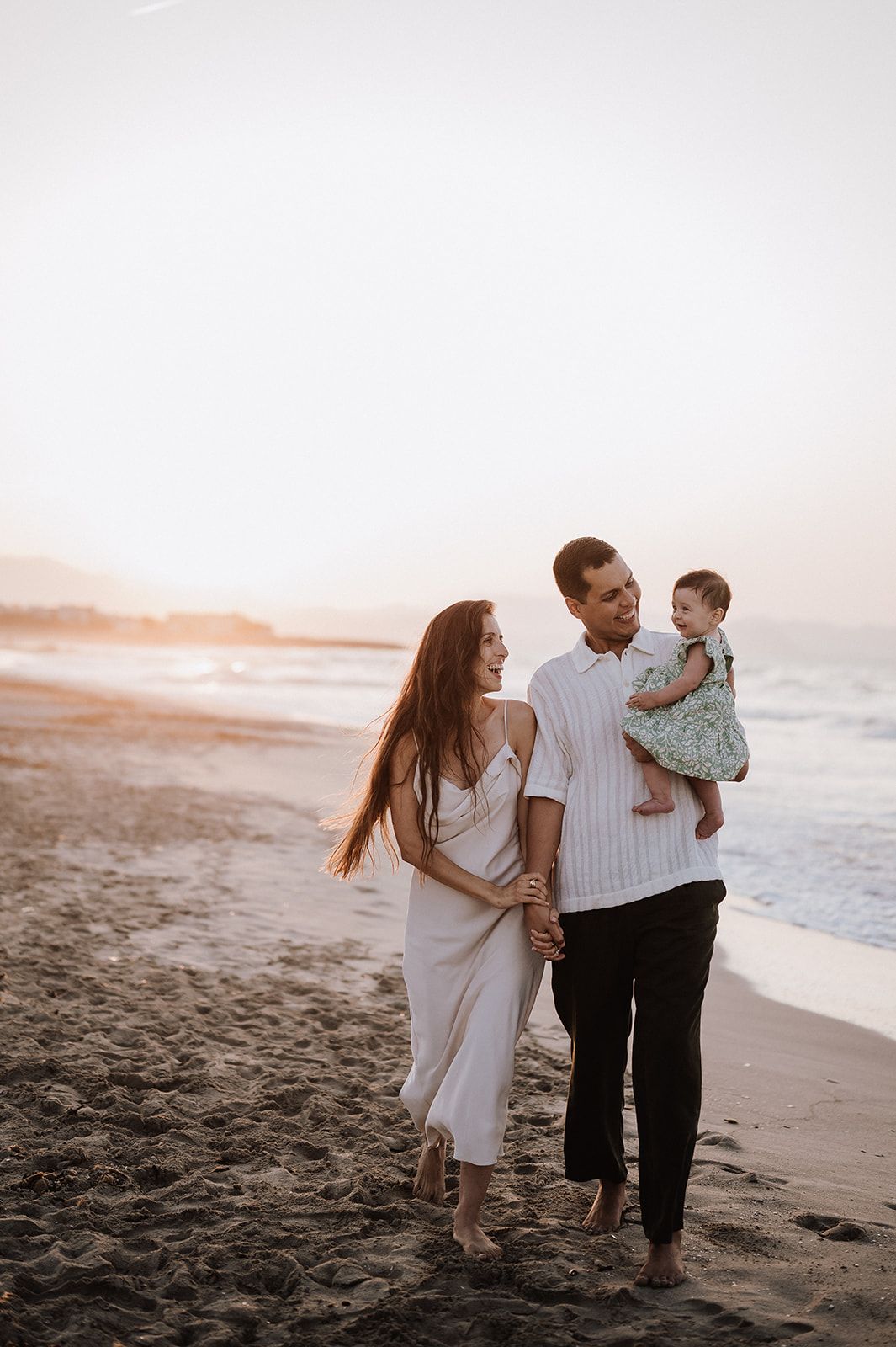 Happy couple walking on beach at sunset, man holding baby girl in green dress near ocean waves.