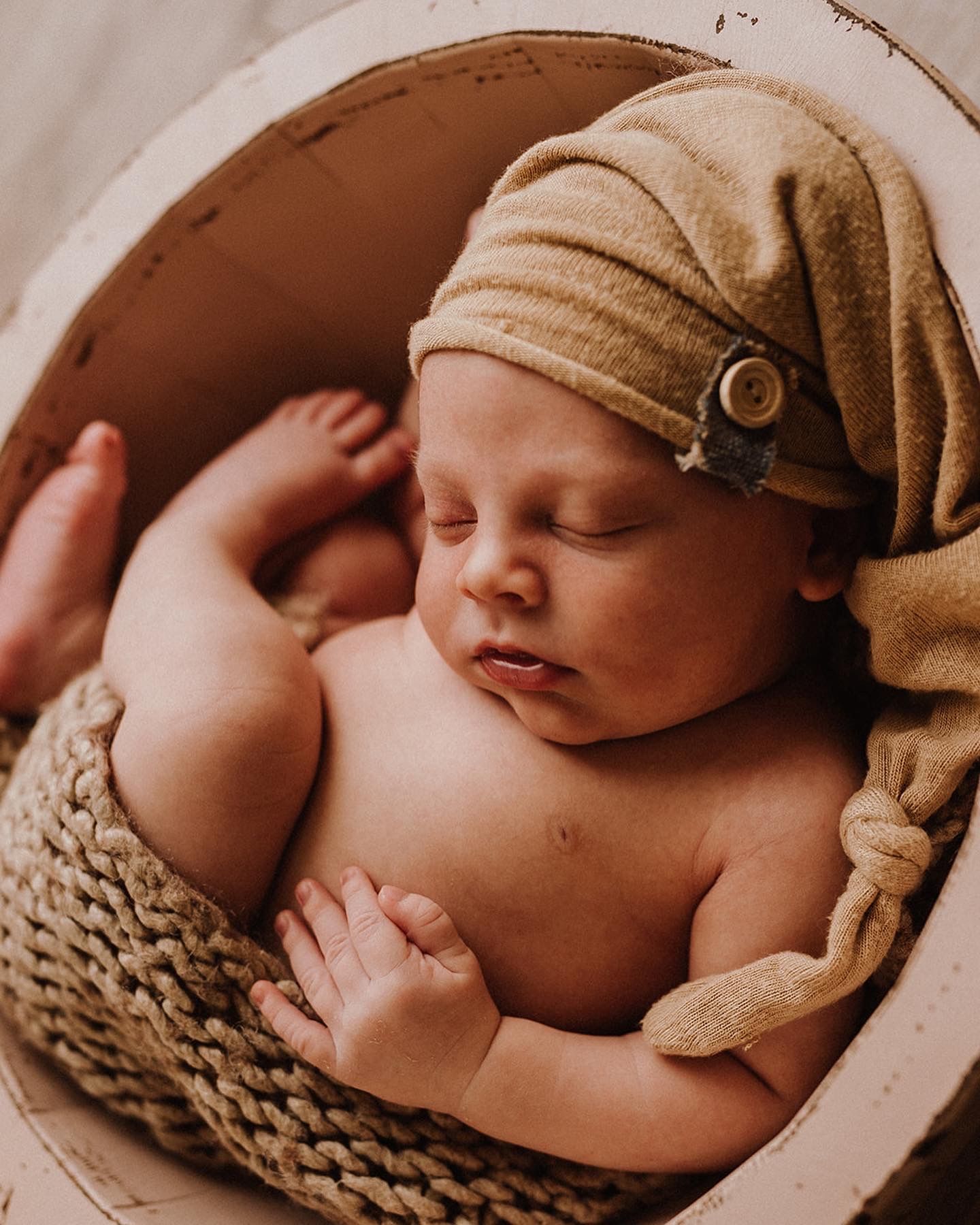 Sleeping newborn baby wearing a tan knit hat, curled up in a round woven basket for a professional newborn photo.