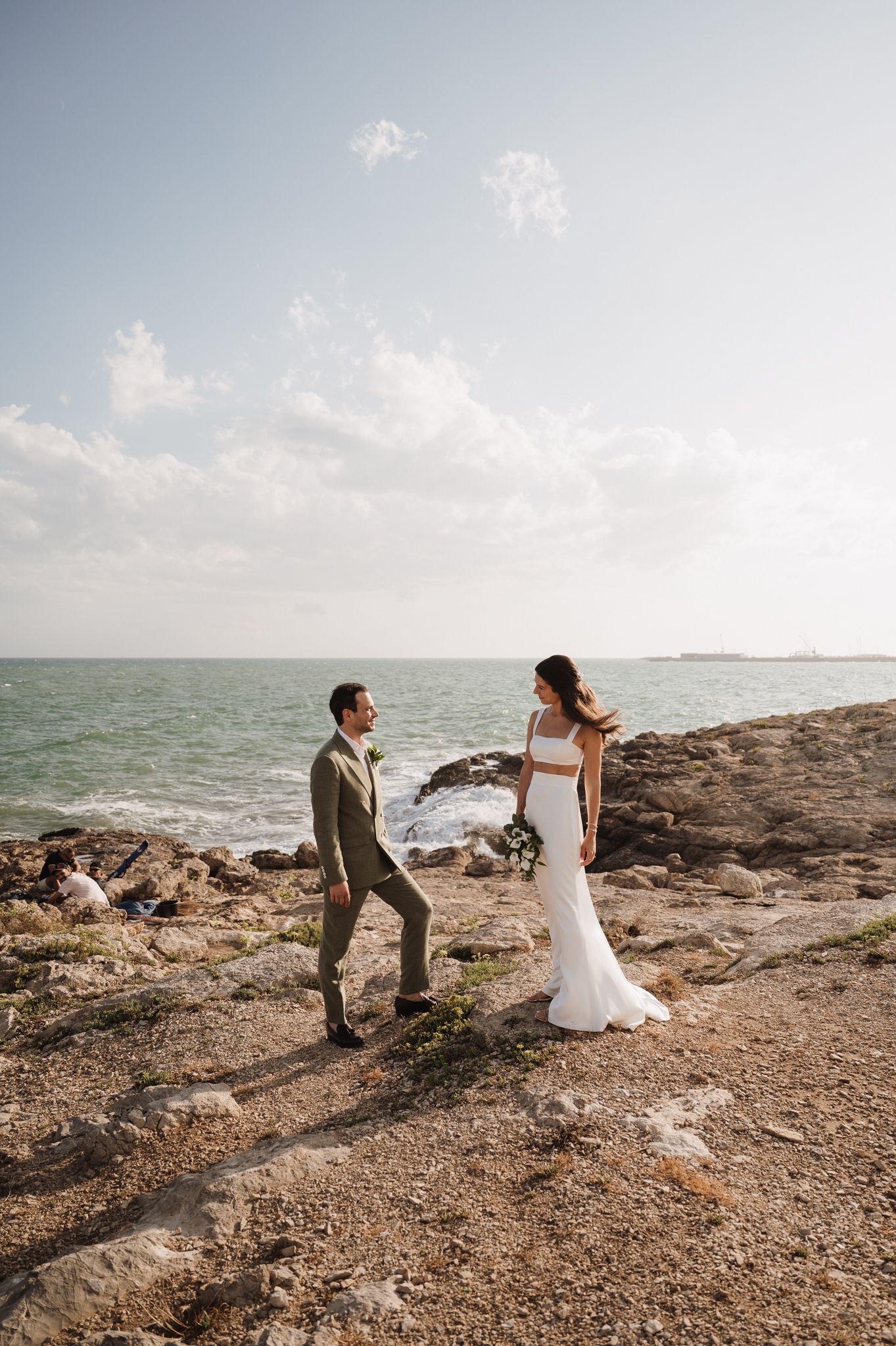 Bride and groom stand on rocky coastal cliffs, ocean waves behind them, she holds a bouquet in a white gown, he wears a tan suit.