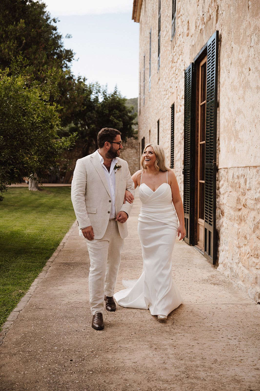 Bride and groom walk hand in hand along stone building path in cream suit and fitted white wedding gown.