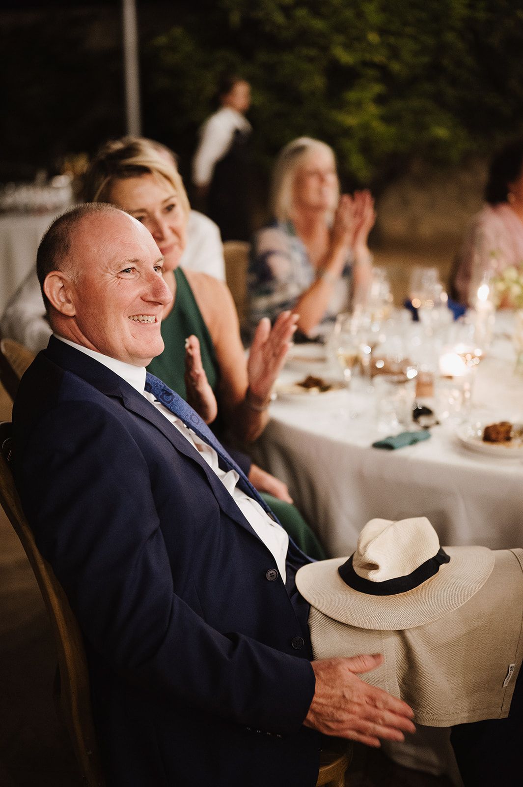 Smiling older gentleman in navy suit applauds at elegant candlelit outdoor wedding reception dinner.