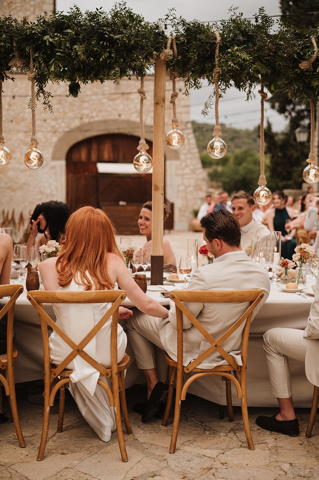Bride and groom seated at outdoor wedding reception table under hanging Edison bulb lights.
