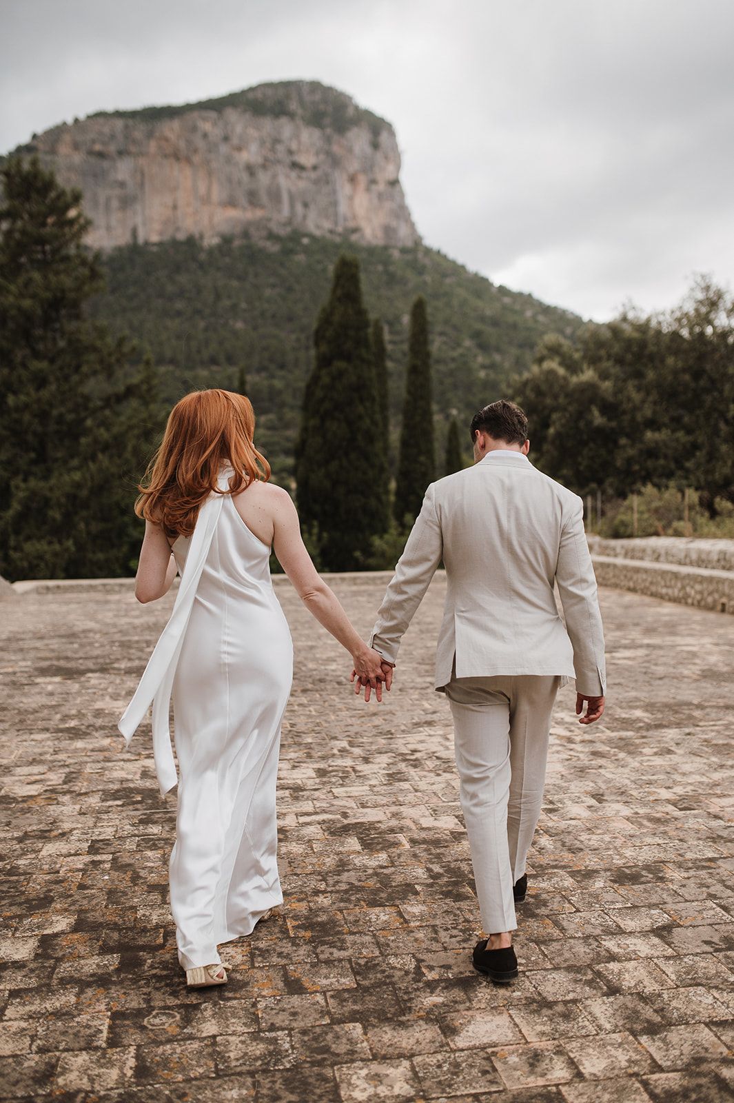 Wedding couple walks away hand in hand on cobblestone path with mountain and cypress trees behind.