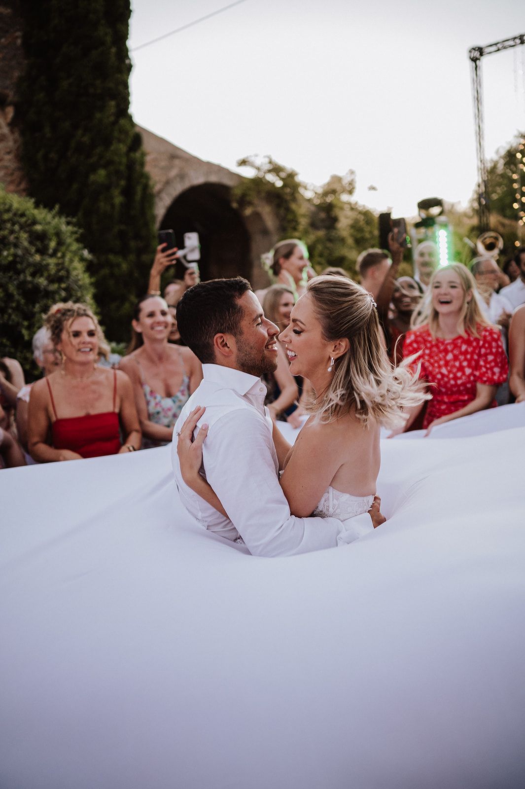 Bride and groom share romantic kiss surrounded by cheering guests at outdoor wedding.