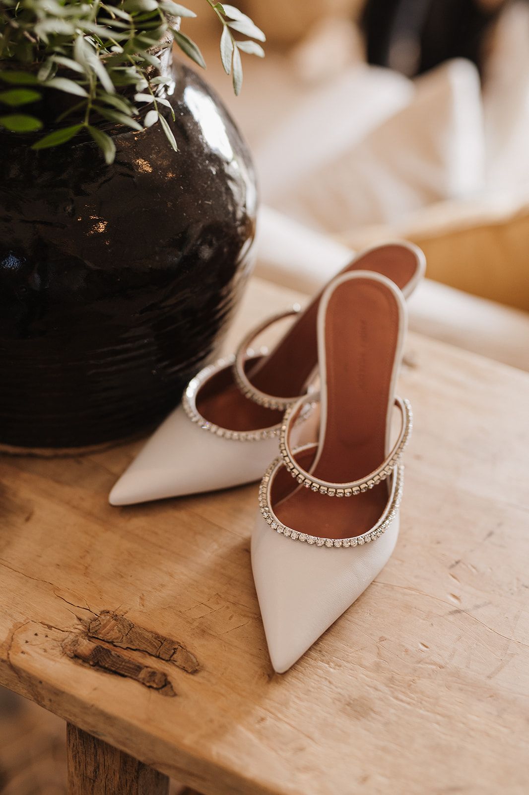 Elegant white pointed-toe bridal heels with crystal embellishments on a rustic wooden table beside a dark ceramic vase.
