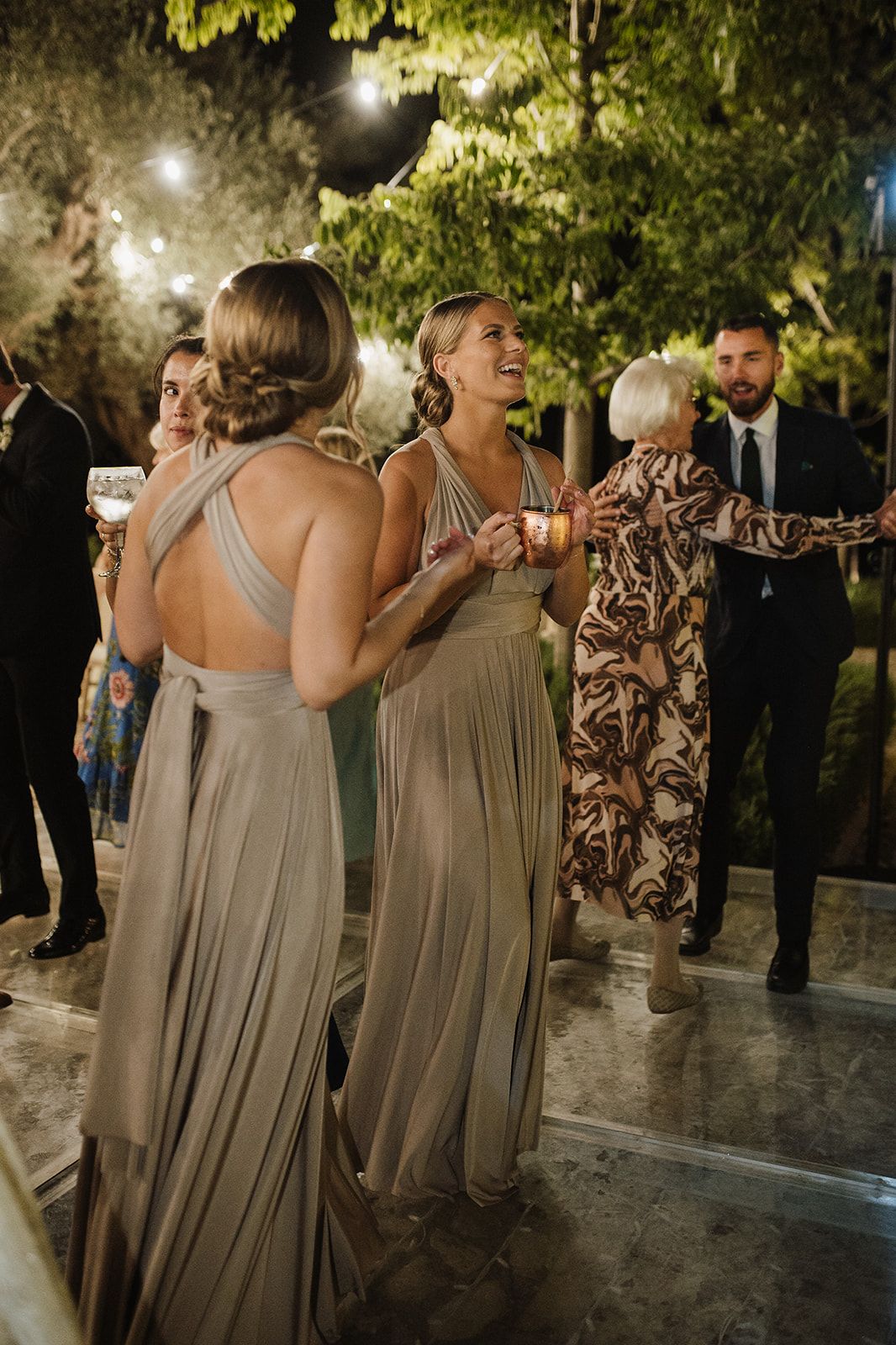 Bridesmaids in elegant taupe gowns laugh and dance at outdoor evening wedding reception.