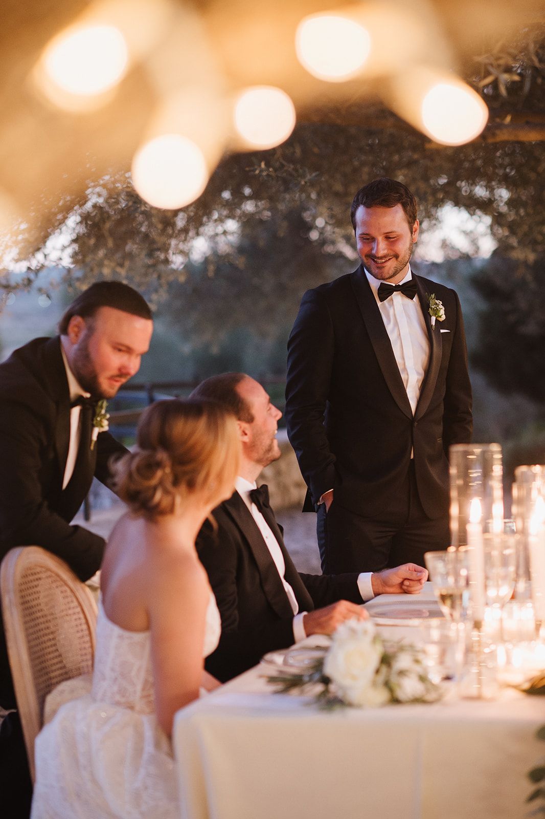 Groomsman delivers toast at candlelit wedding reception table as bride and groom smile warmly under string lights.