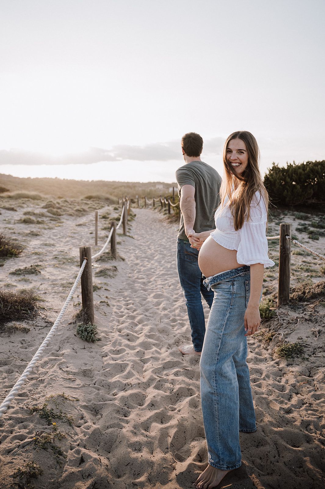 Pregnant woman in white top and jeans smiles on sandy beach path at sunset with partner walking ahead.