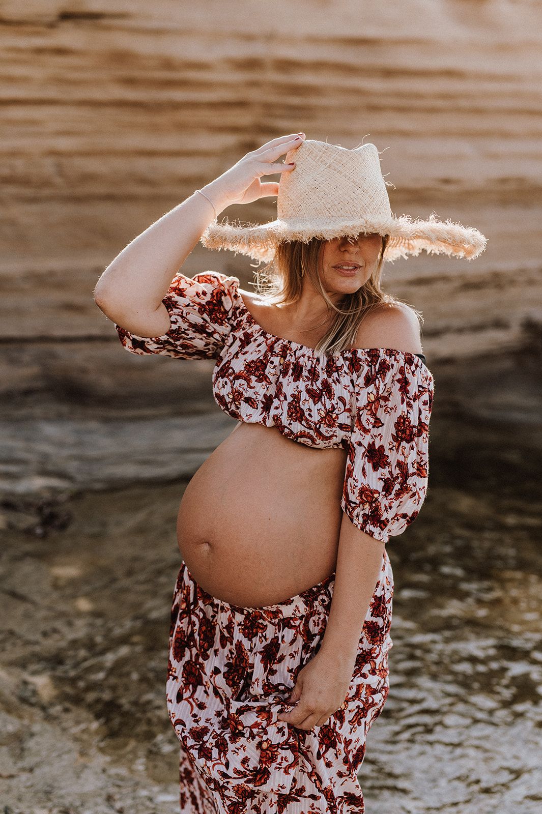 Pregnant woman in floral crop top and maxi skirt holding straw hat at golden hour beach.