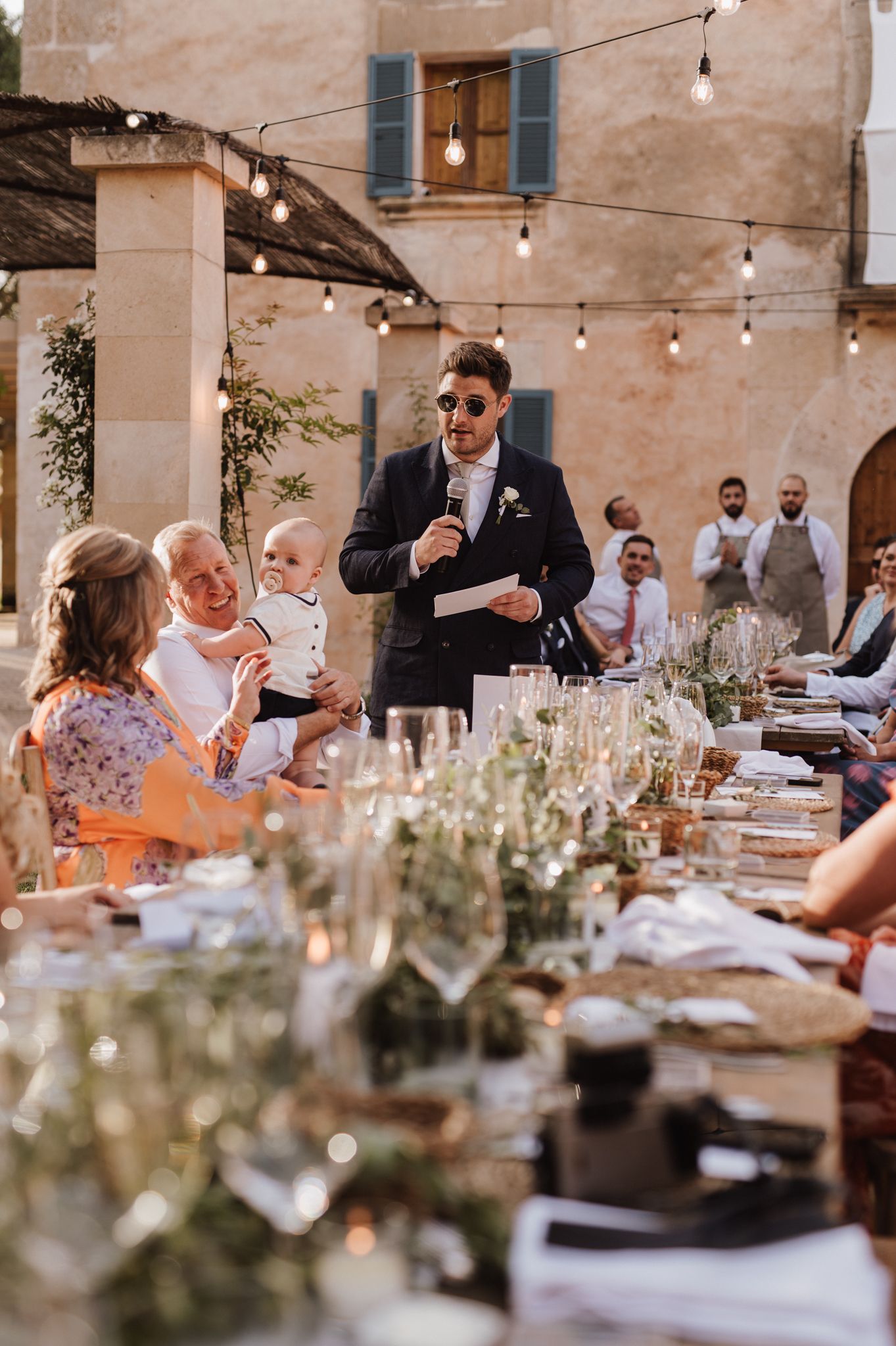 Groom delivering speech at elegant outdoor wedding reception with long decorated table.