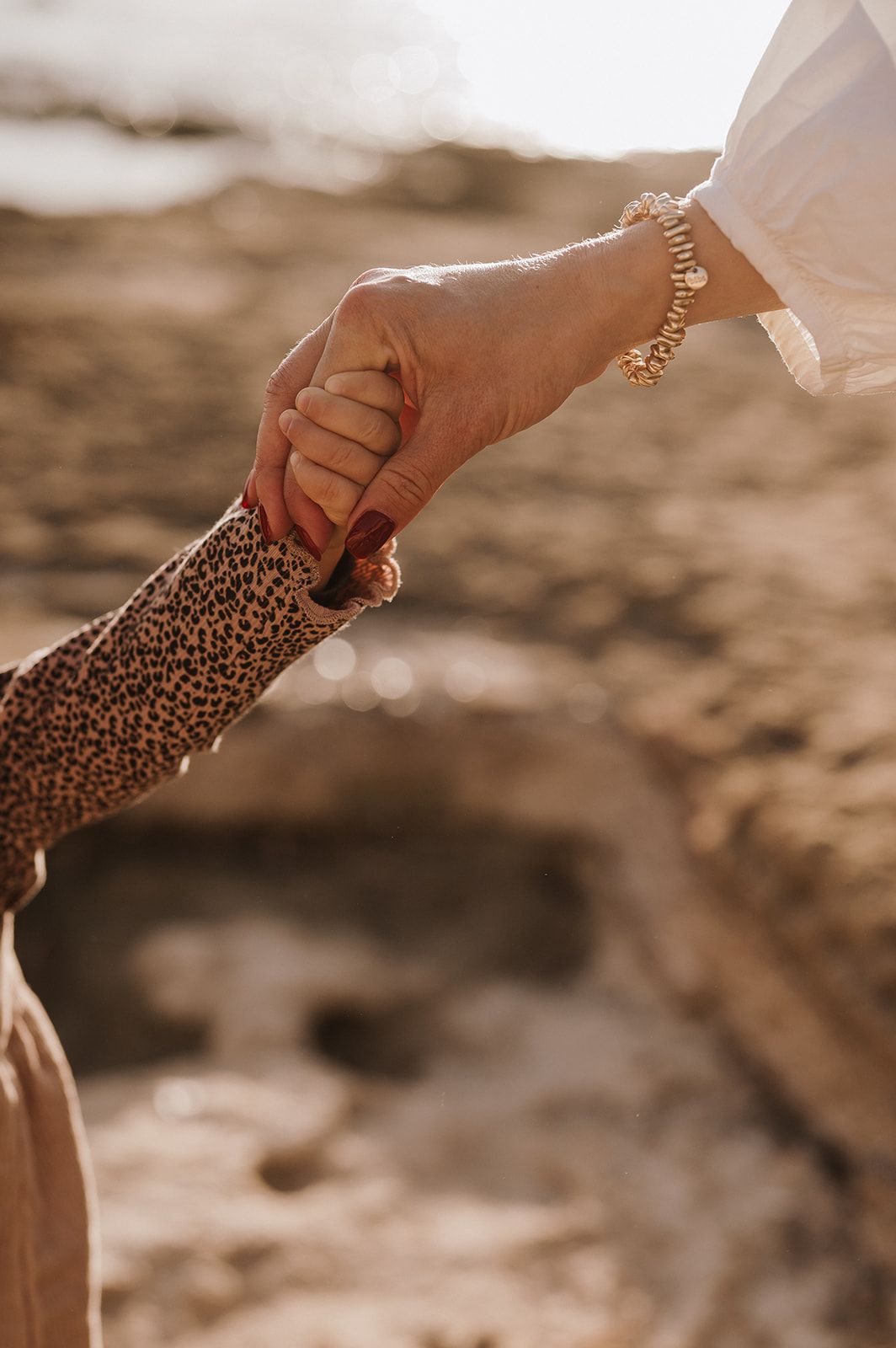 Close-up of adult and child holding hands at the beach, woman wearing gold bracelet in warm sunlight.